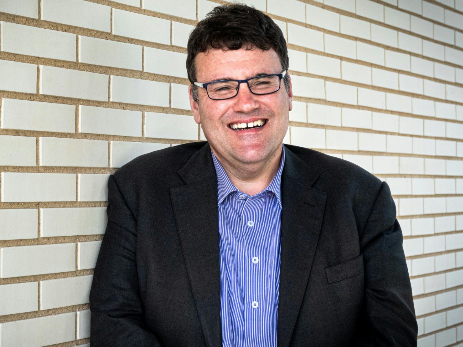 A man with glasses wearing a sttriped button down shirt and black blazer, standing in front of a whi