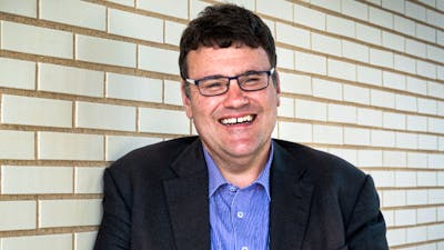 A man with glasses wearing a sttriped button down shirt and black blazer, standing in front of a whi