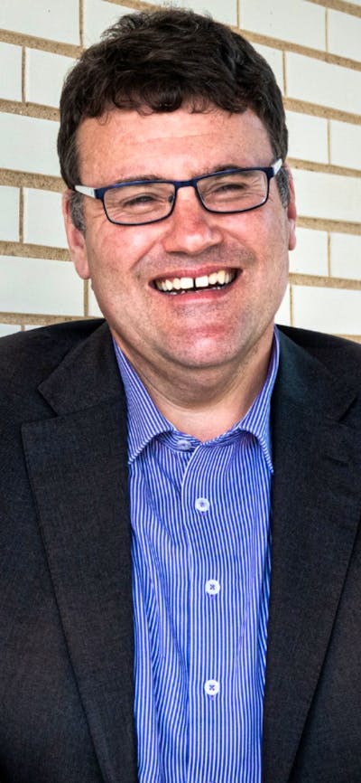 A man with glasses wearing a sttriped button down shirt and black blazer, standing in front of a whi