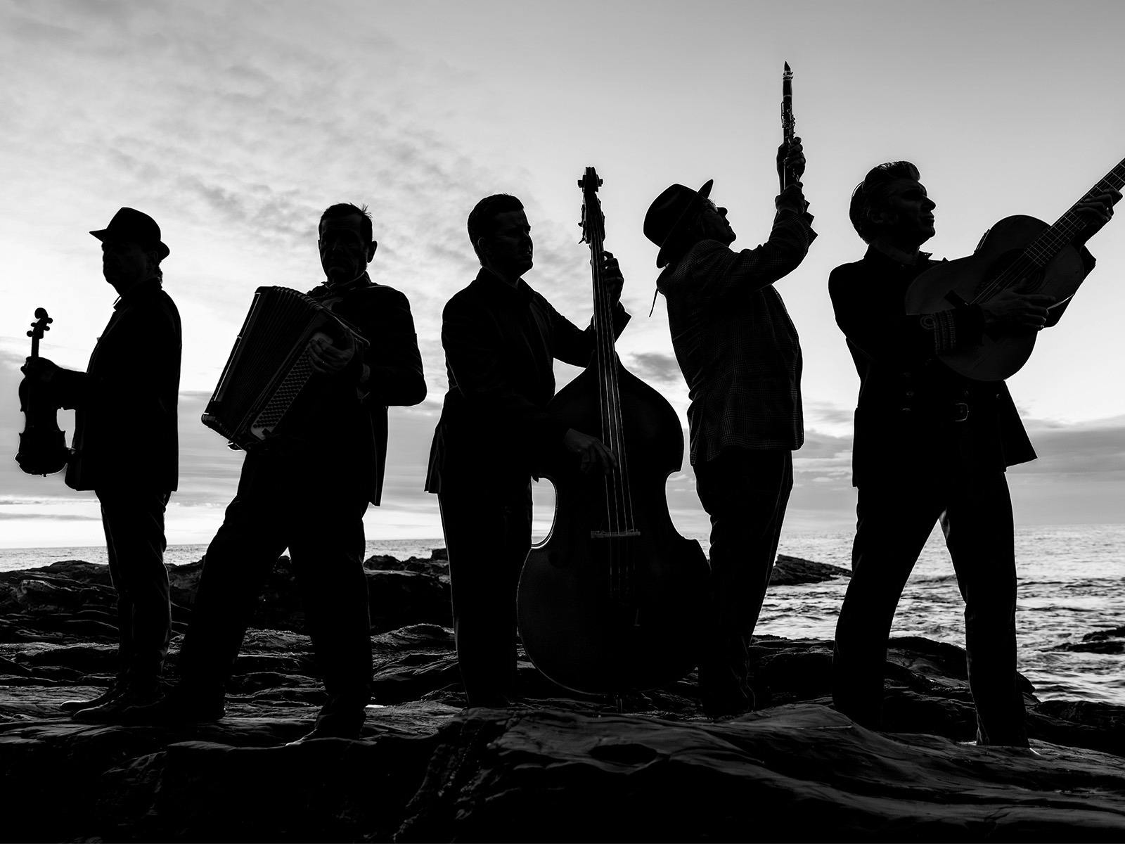 Black-and-white photograph of five silhouetted musicians performing on rocky shore at sunset, playin