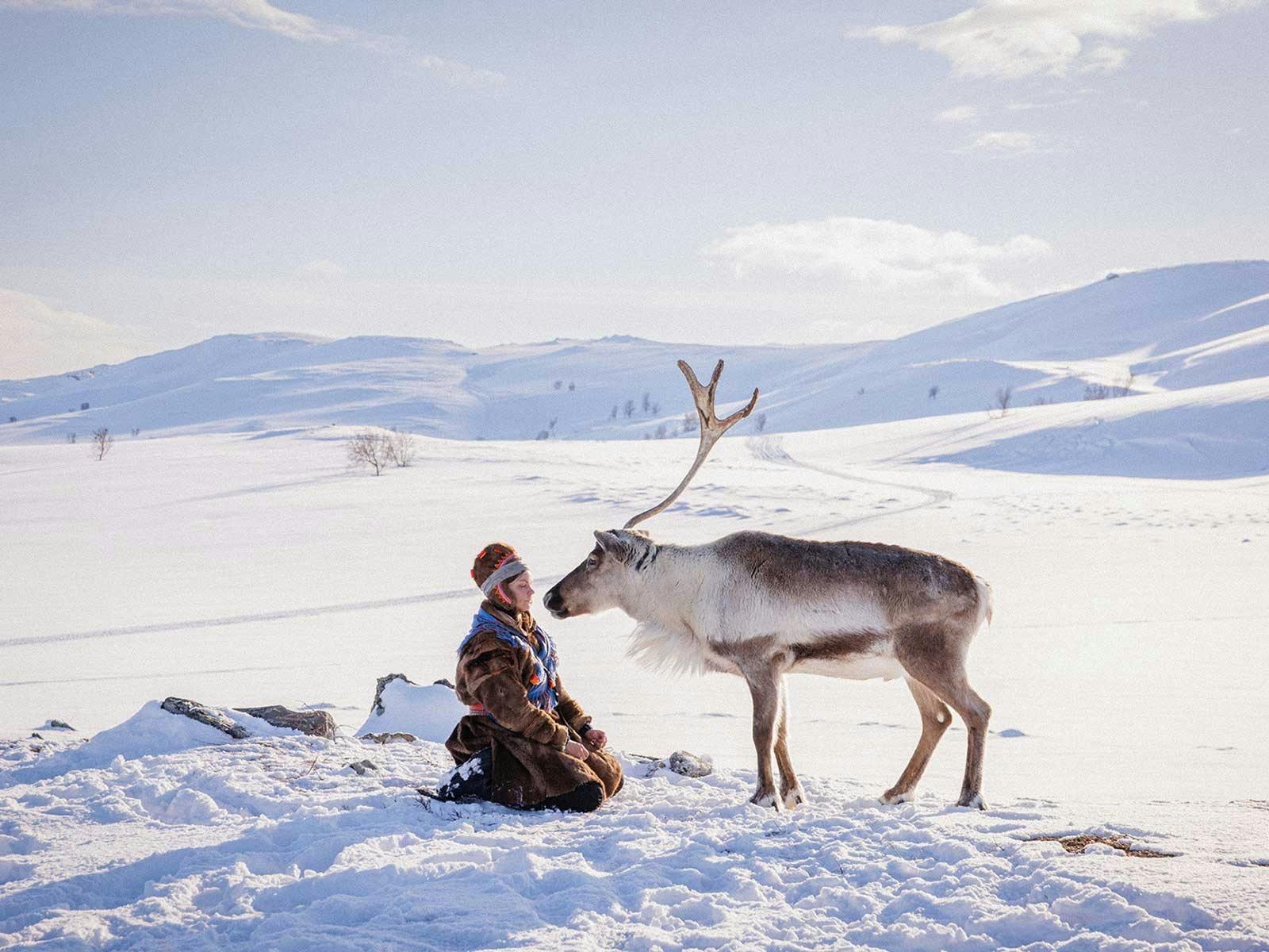 A woman crouches in the snow greeting a large reindeer