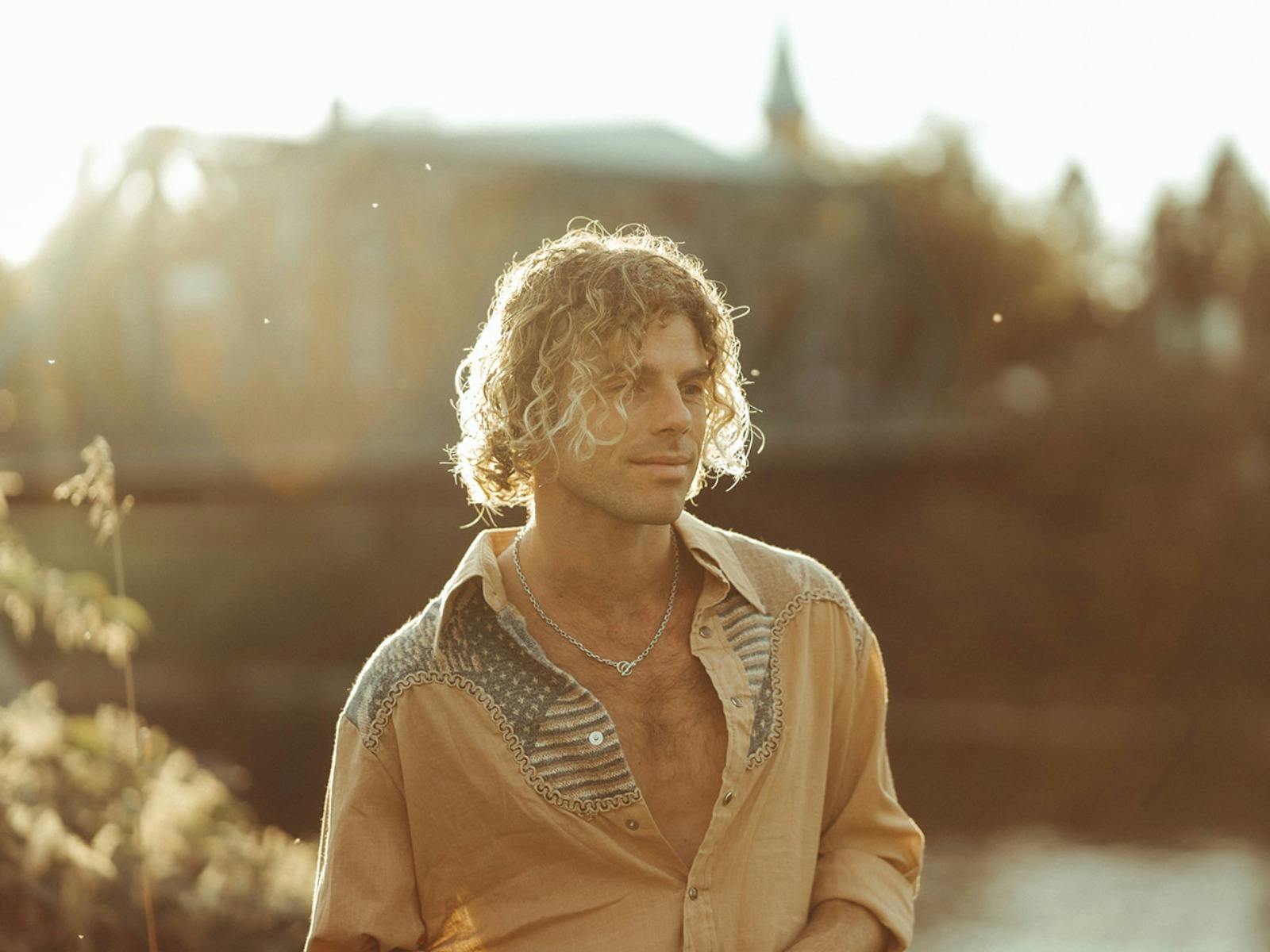 Man with blonde curly shoulder length hair standing with afternoon sunlight on him