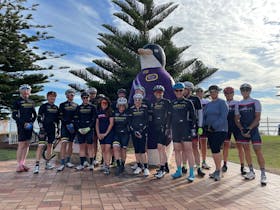Group of cyclists standing in front of a large statue of a penguin on the main street of Penguin Tas
