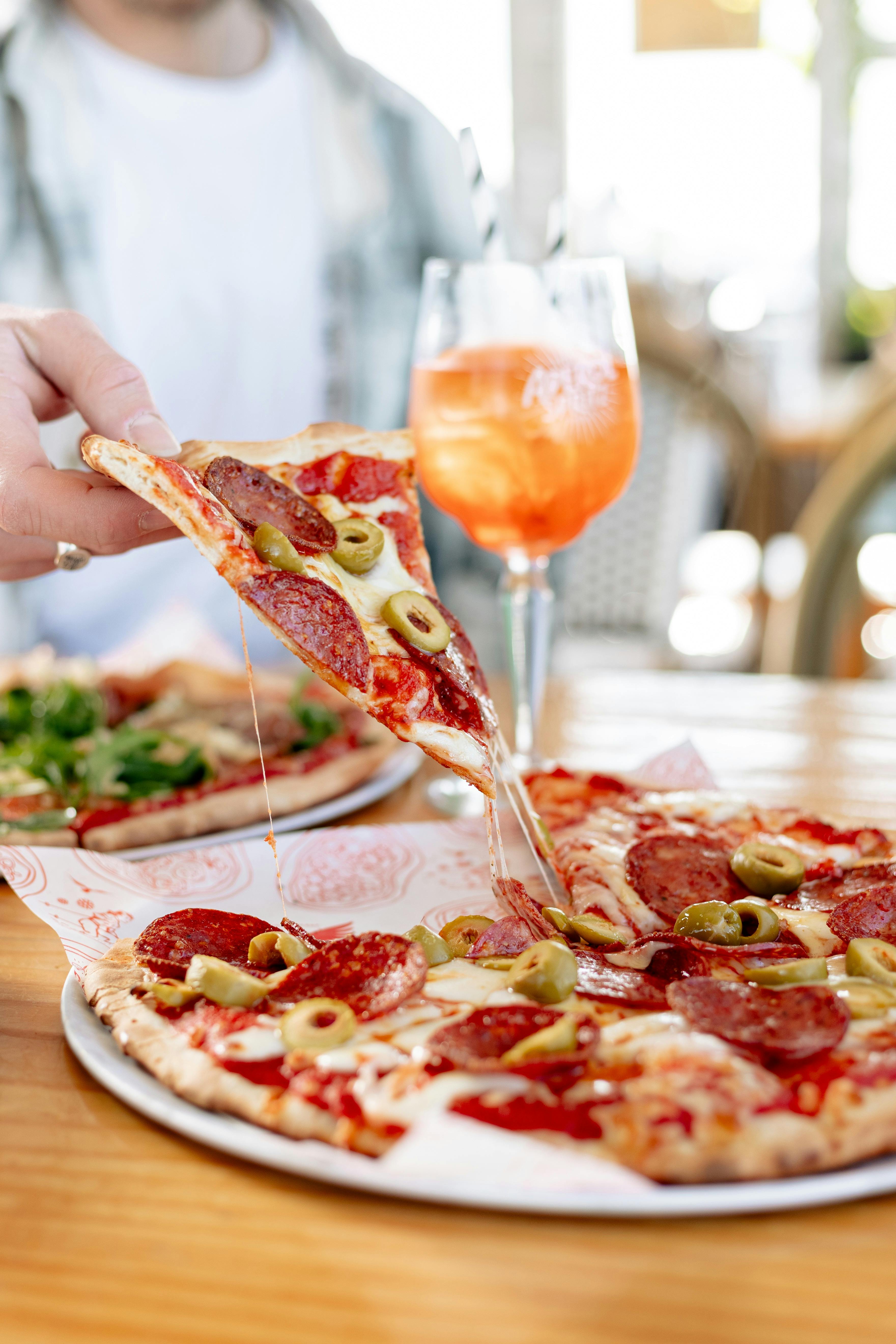 A slice of Pepperoni pizza being lifted from the tray with an Aperol spritz in the background