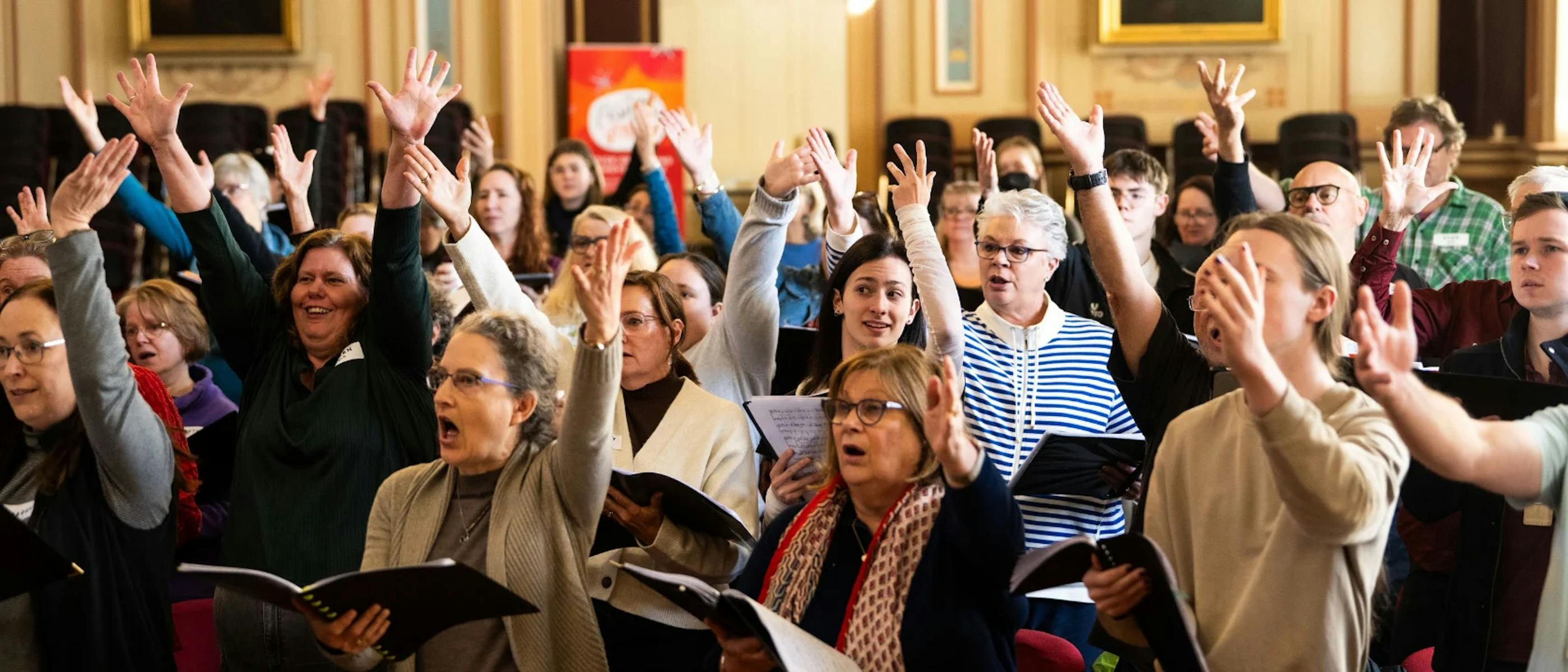A group of singers smiling and singing together holding songbooks with their arms up