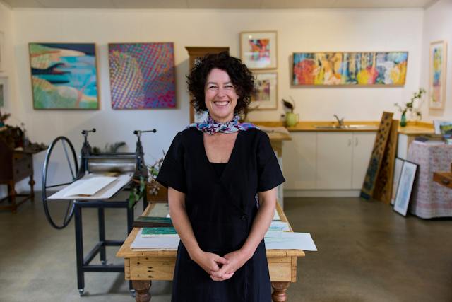 Smiling lady in black standing in front of colourful art and a printing press