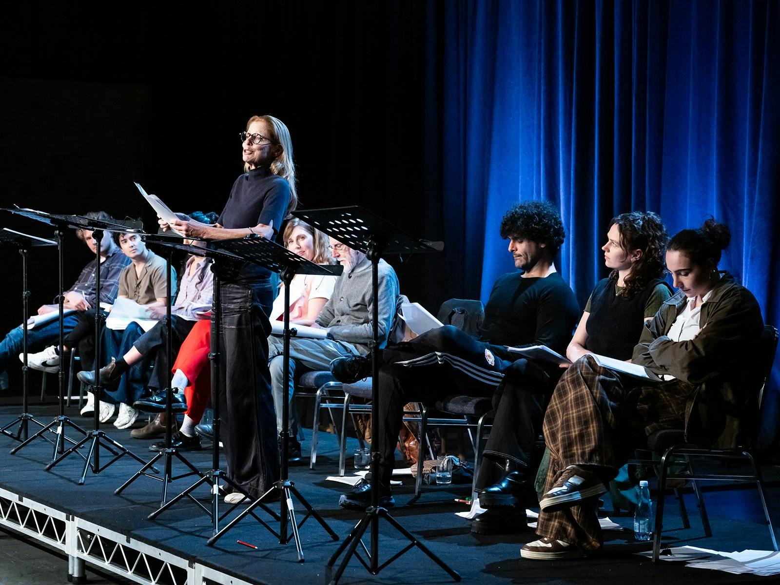 A group of actors are sitting and standing behind a row of music stands