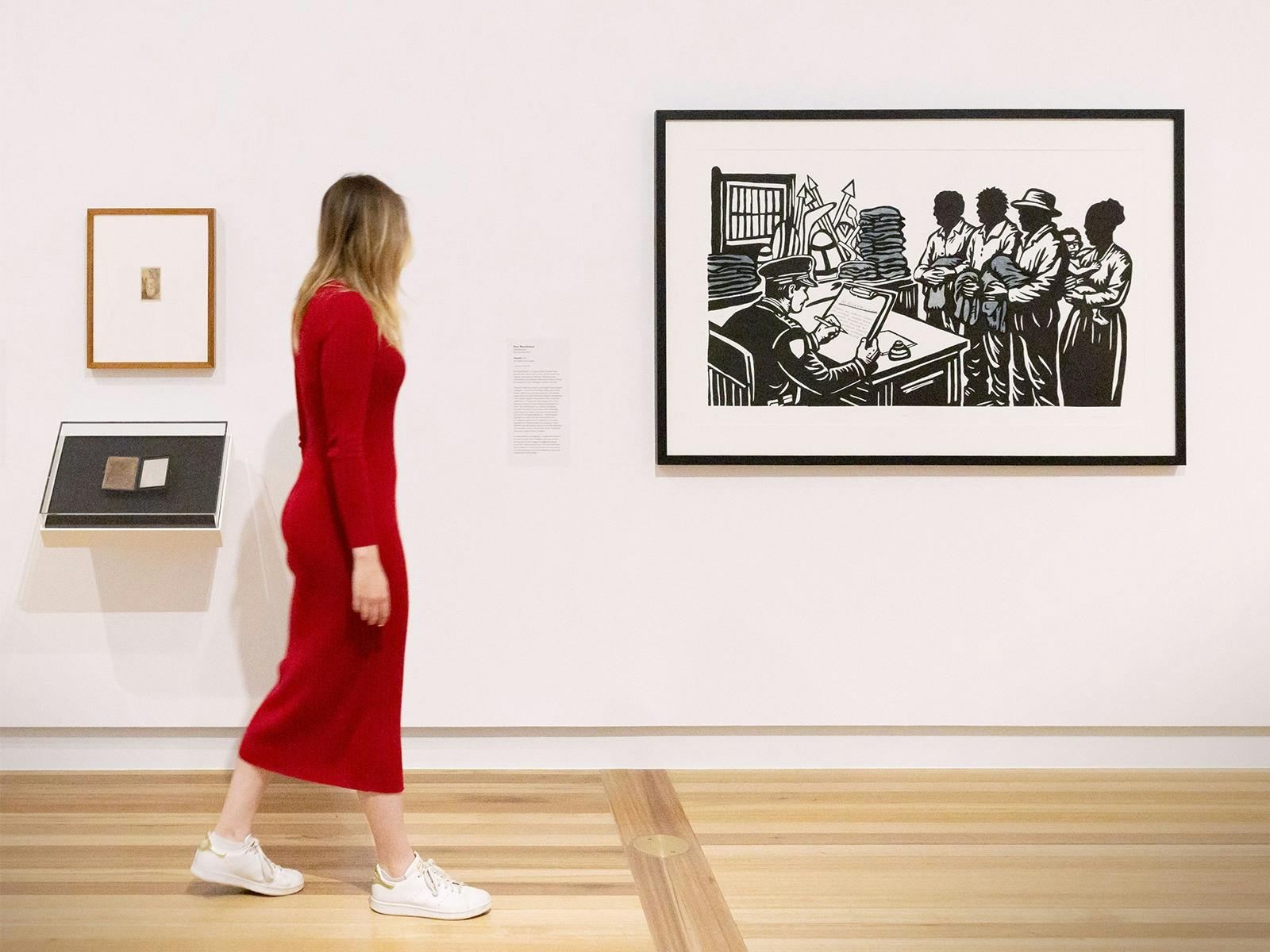 A person wearing a pink dress walks by a Gallery wall, looking at several artworks.