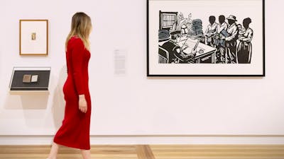 A person wearing a pink dress walks by a Gallery wall, looking at several artworks.