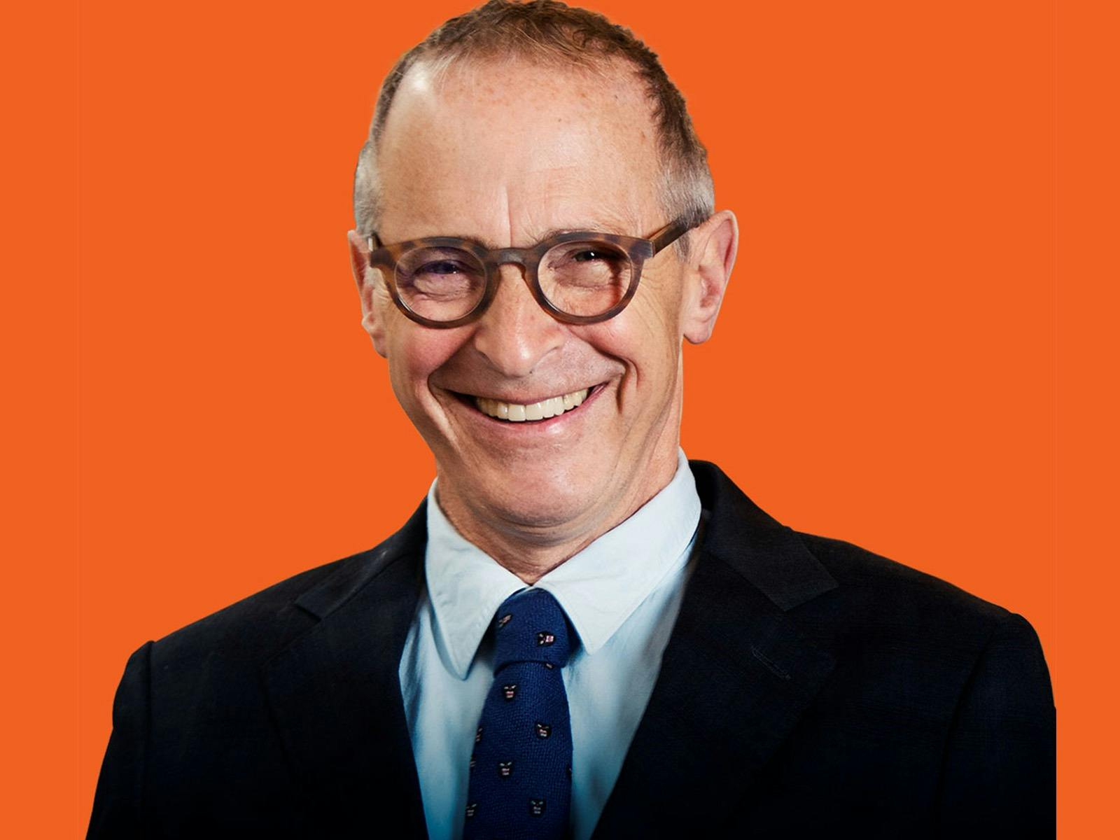 A smiling middle-aged man with short brown hair, round dark-framed glasses, and a dark suit jacket,