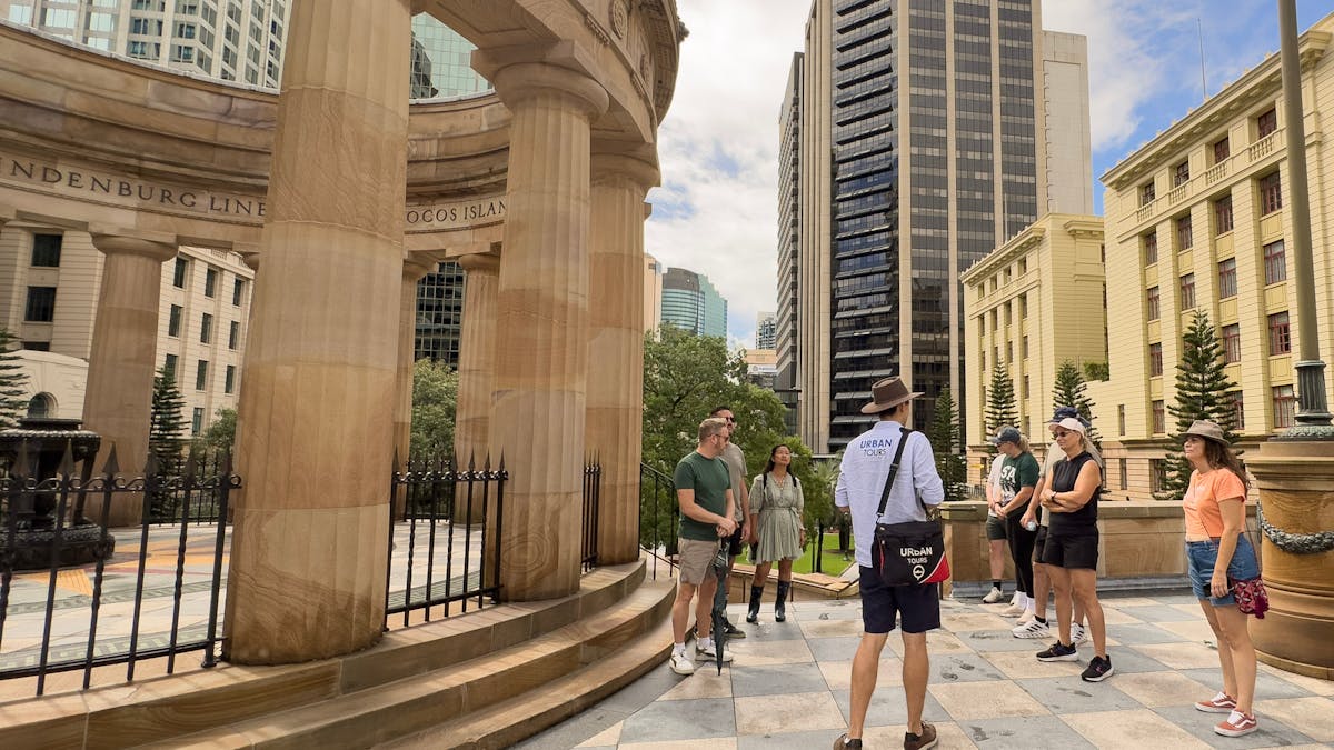 Tour guide standing next to columns of a shrine talking to a group of people.  Buildings in the back