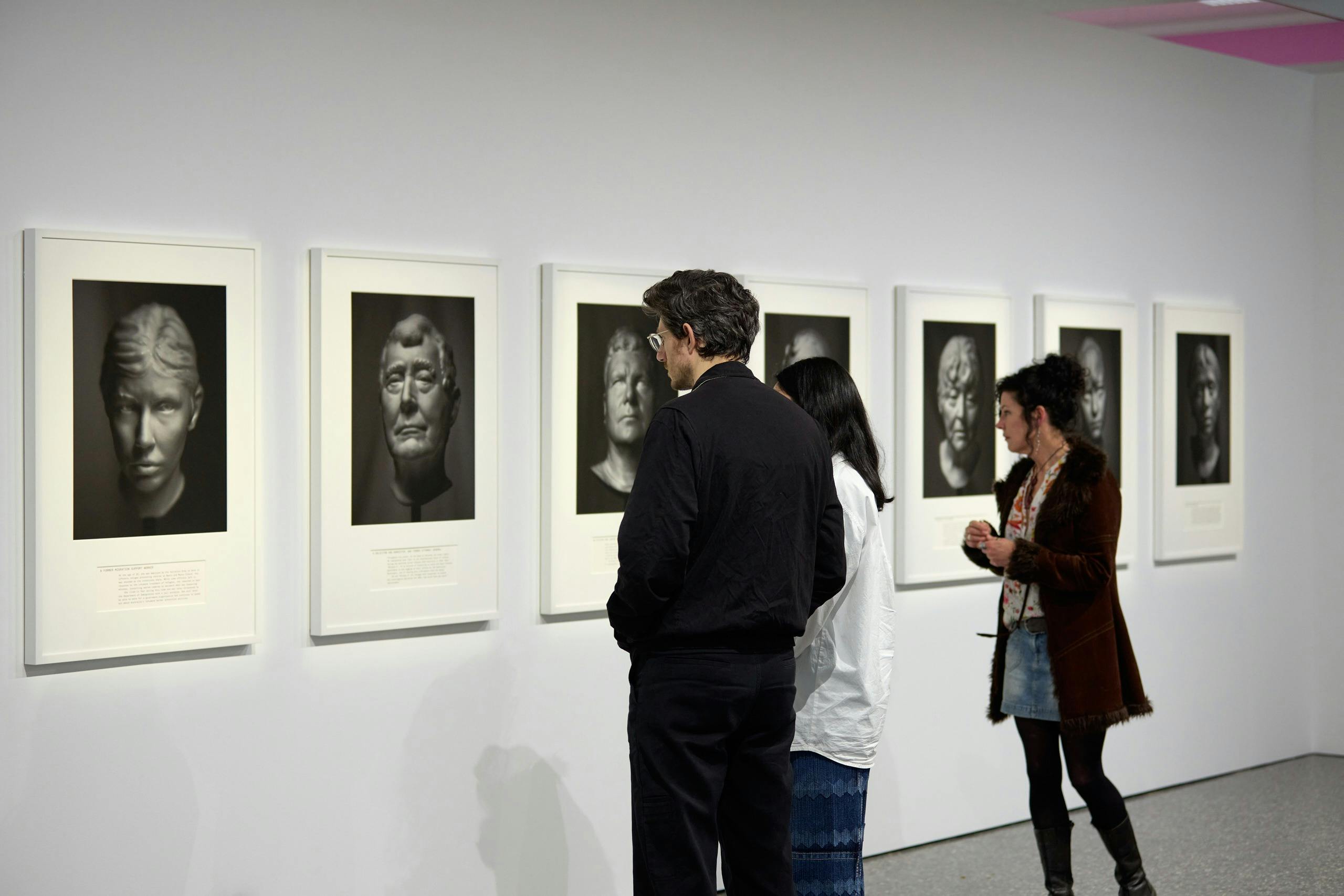 Five blurred portraits hang on a wall, three people  inspects the artworks