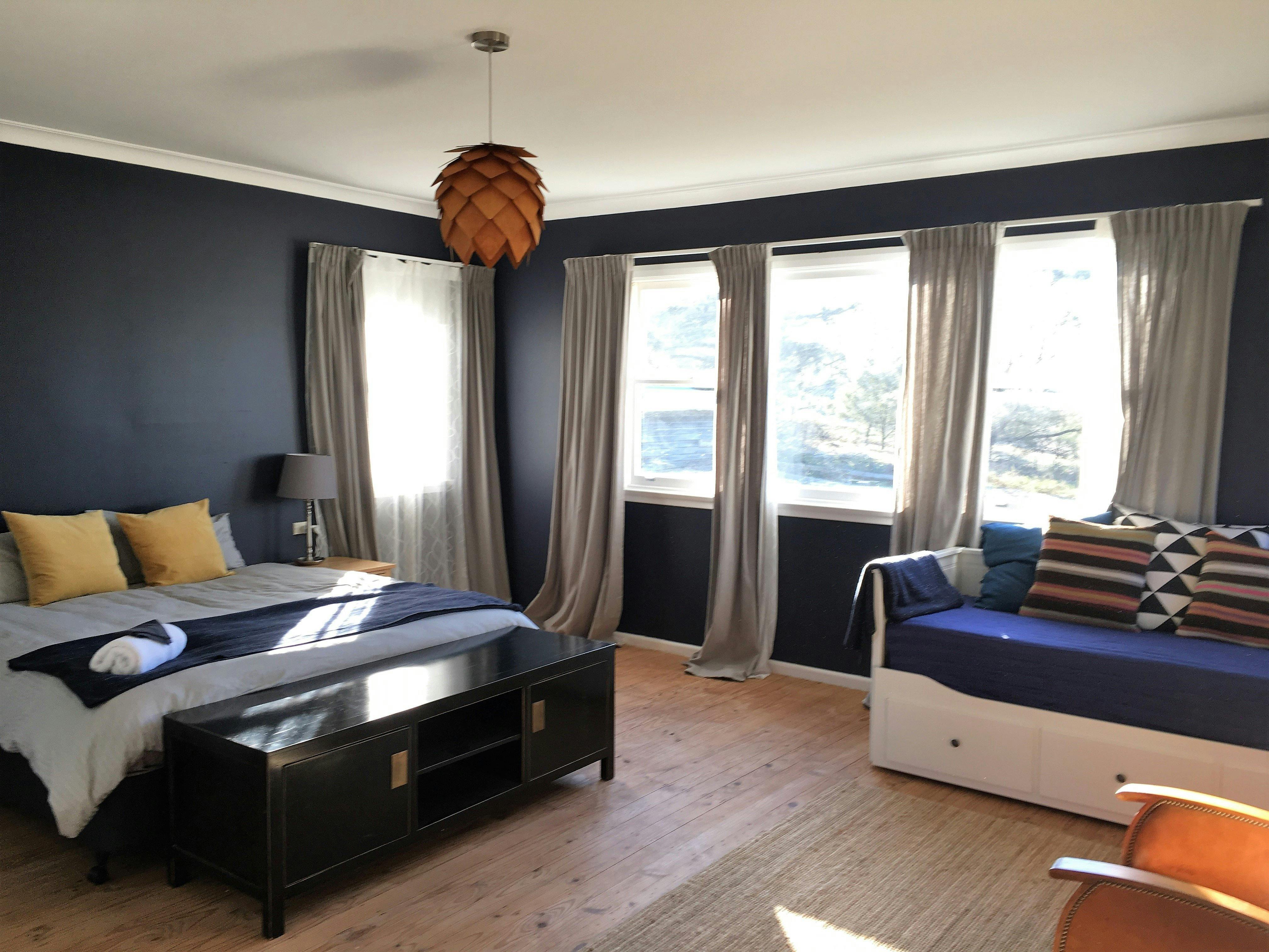 Big sunny bedroom with dark blue walls & light timber floor. King bed & daybed, colourful cushions.