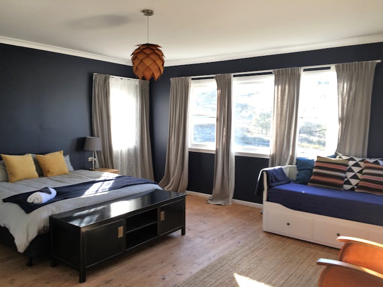 Big sunny bedroom with dark blue walls & light timber floor. King bed & daybed, colourful cushions.