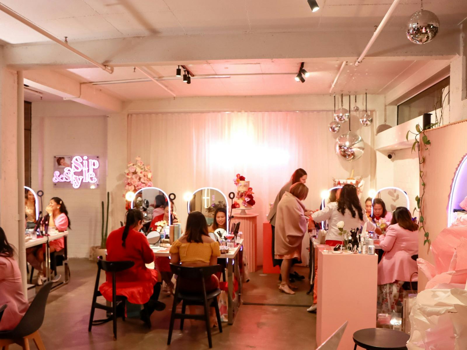 Guests at a makeup masterclass in a pink-lit studio with mirrors, flowers, and disco balls overhead.