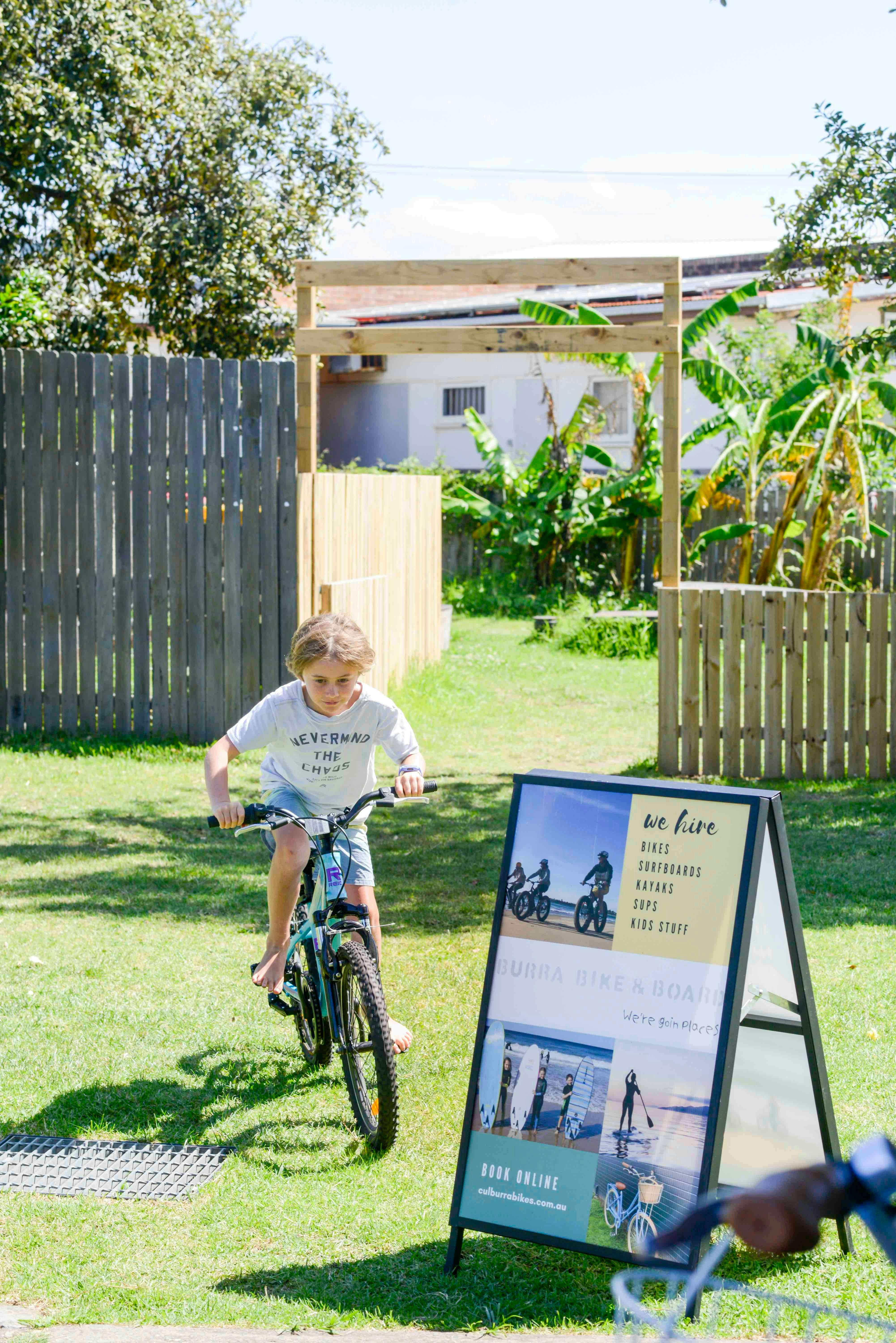 Young boy riding BMX bike through garden, next to signage for Burra Bike & Board surf hire operation