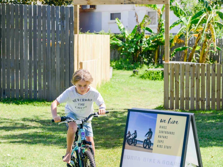 Young boy riding BMX bike through garden, next to signage for Burra Bike & Board surf hire operation