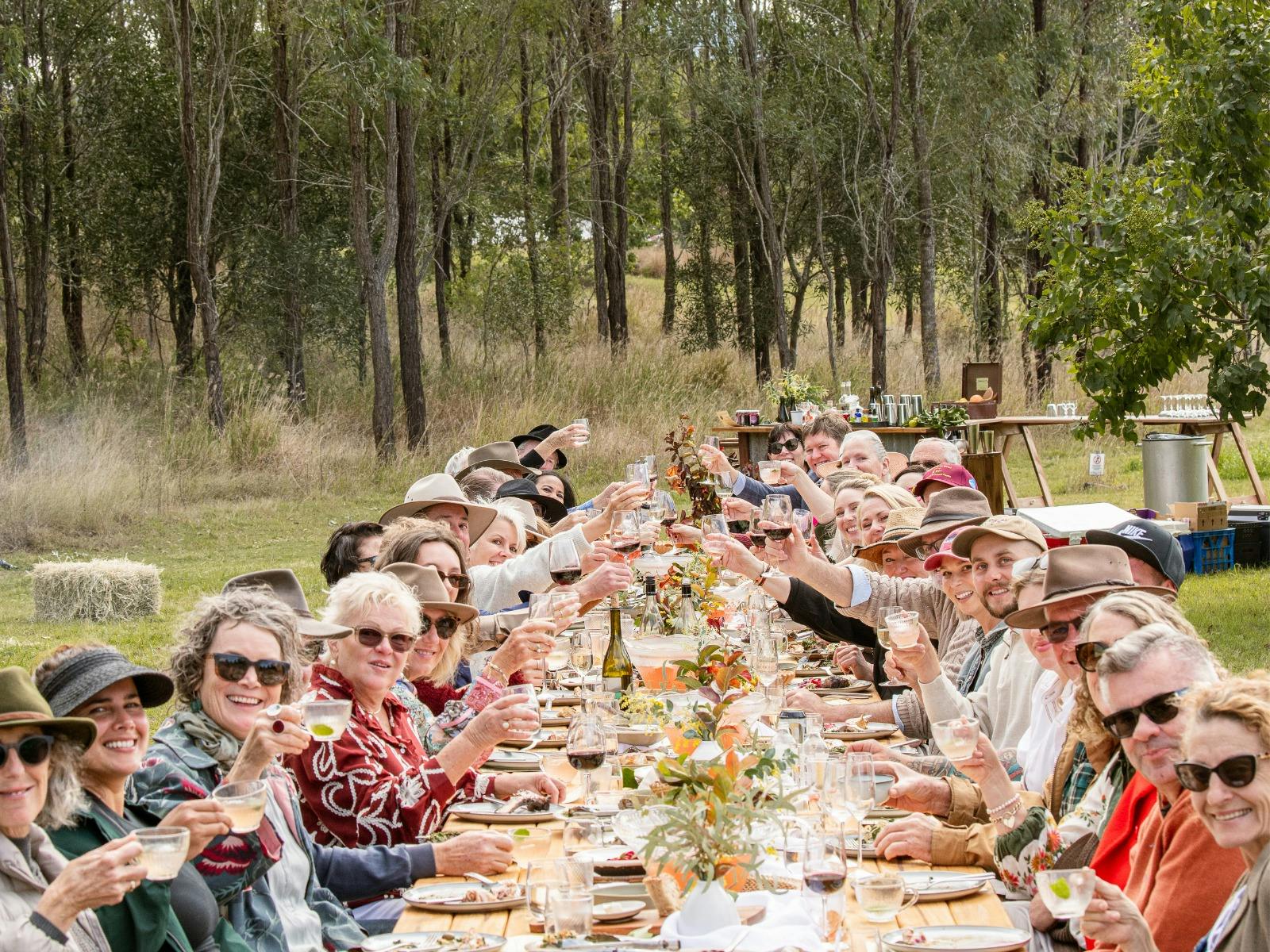 Guests seated at long table lunch all cheering their drinks together and smiling towards the camera