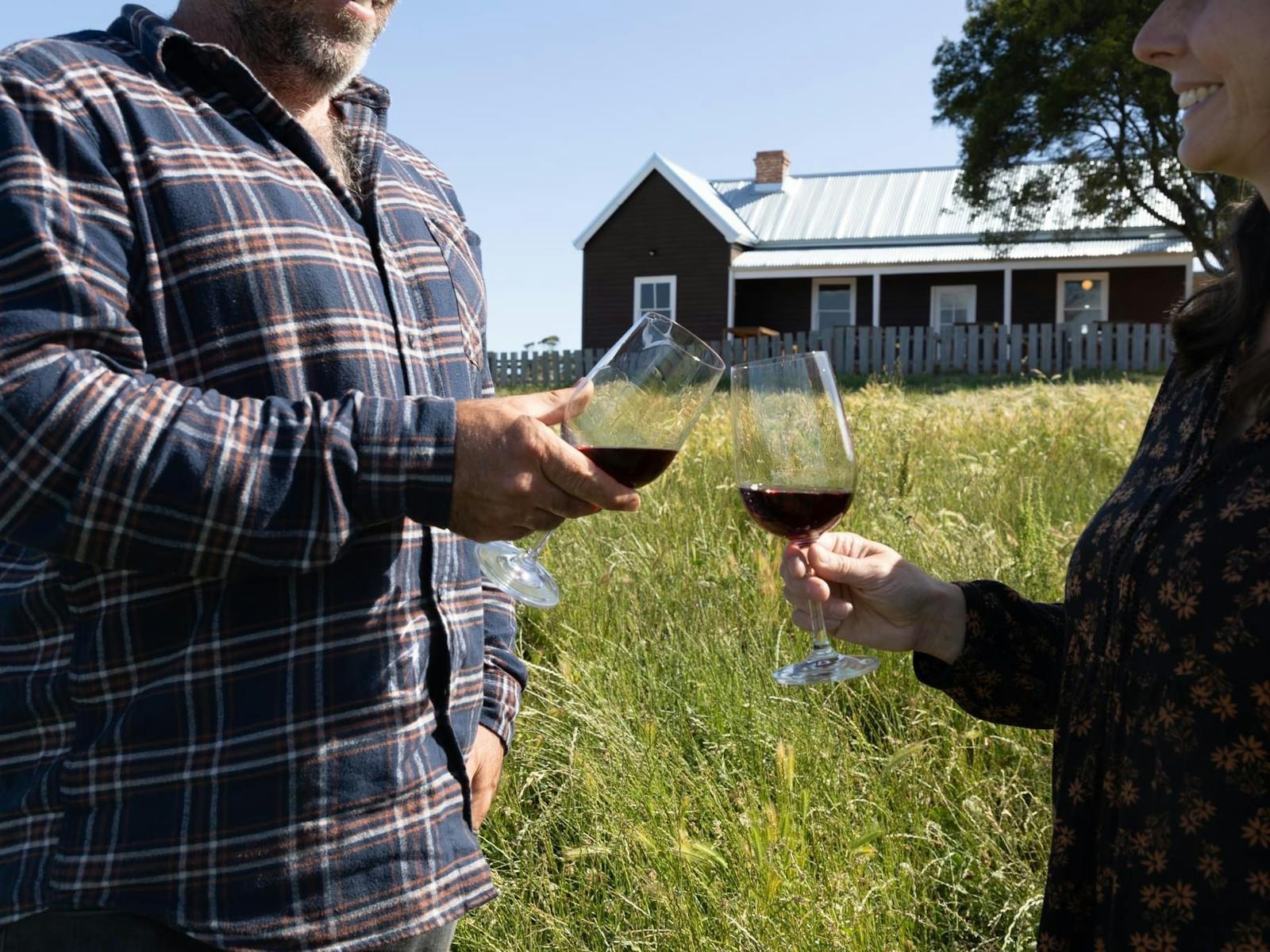 Couple toasting red wine glasses in paddock with brown weatherboard cottage in background.