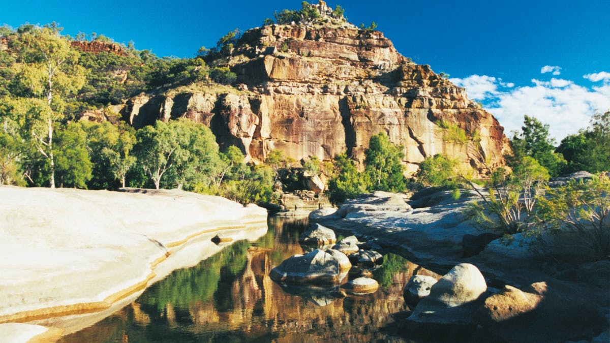 A pyramid-like sandstone formation towers over a small creek with boulders, fringed by vegetation.