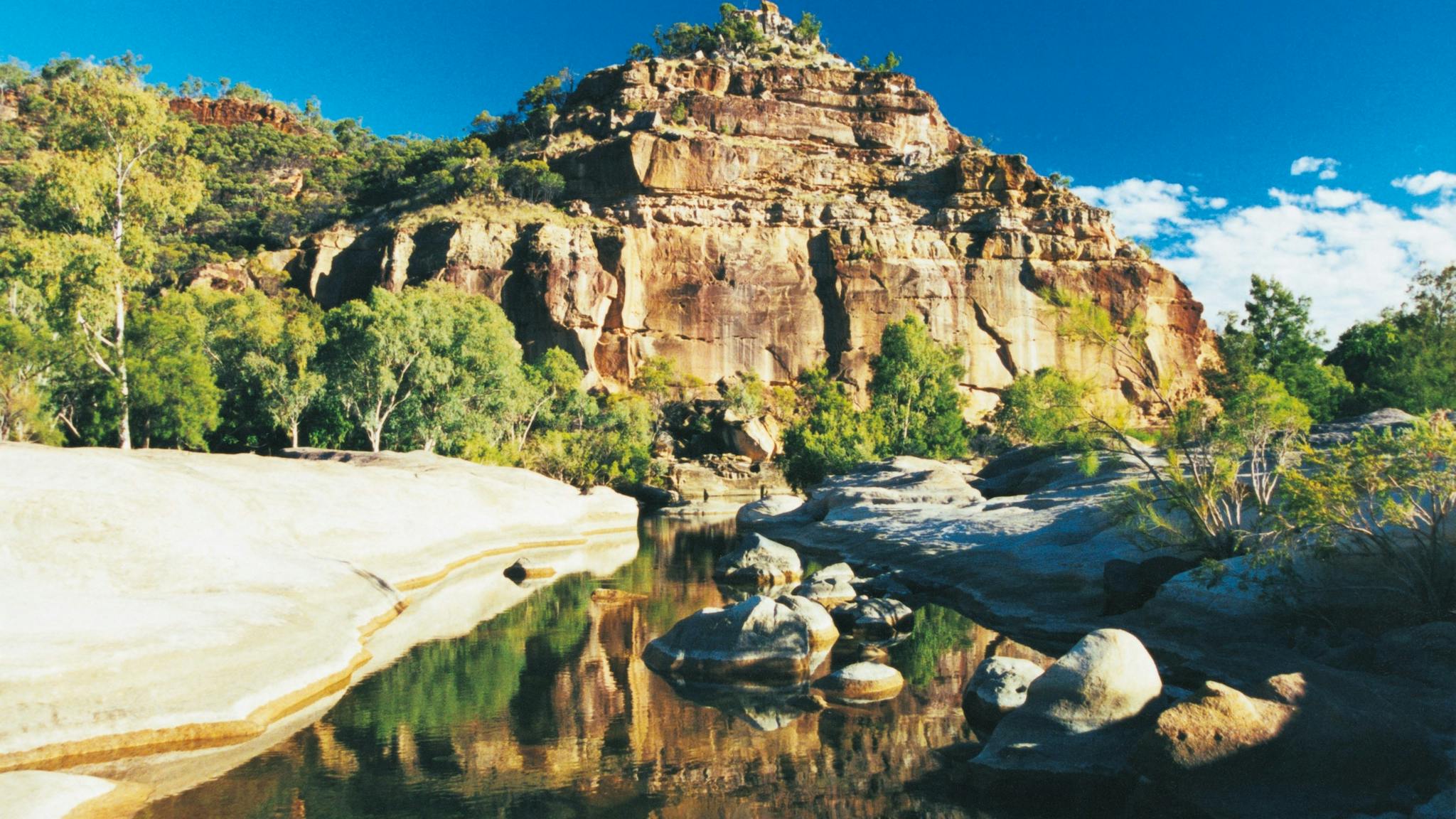 A pyramid-like sandstone formation towers over a small creek with boulders, fringed by vegetation. A pyramid-like sandstone formation towers over a small creek with boulders, fringed by vegetation.