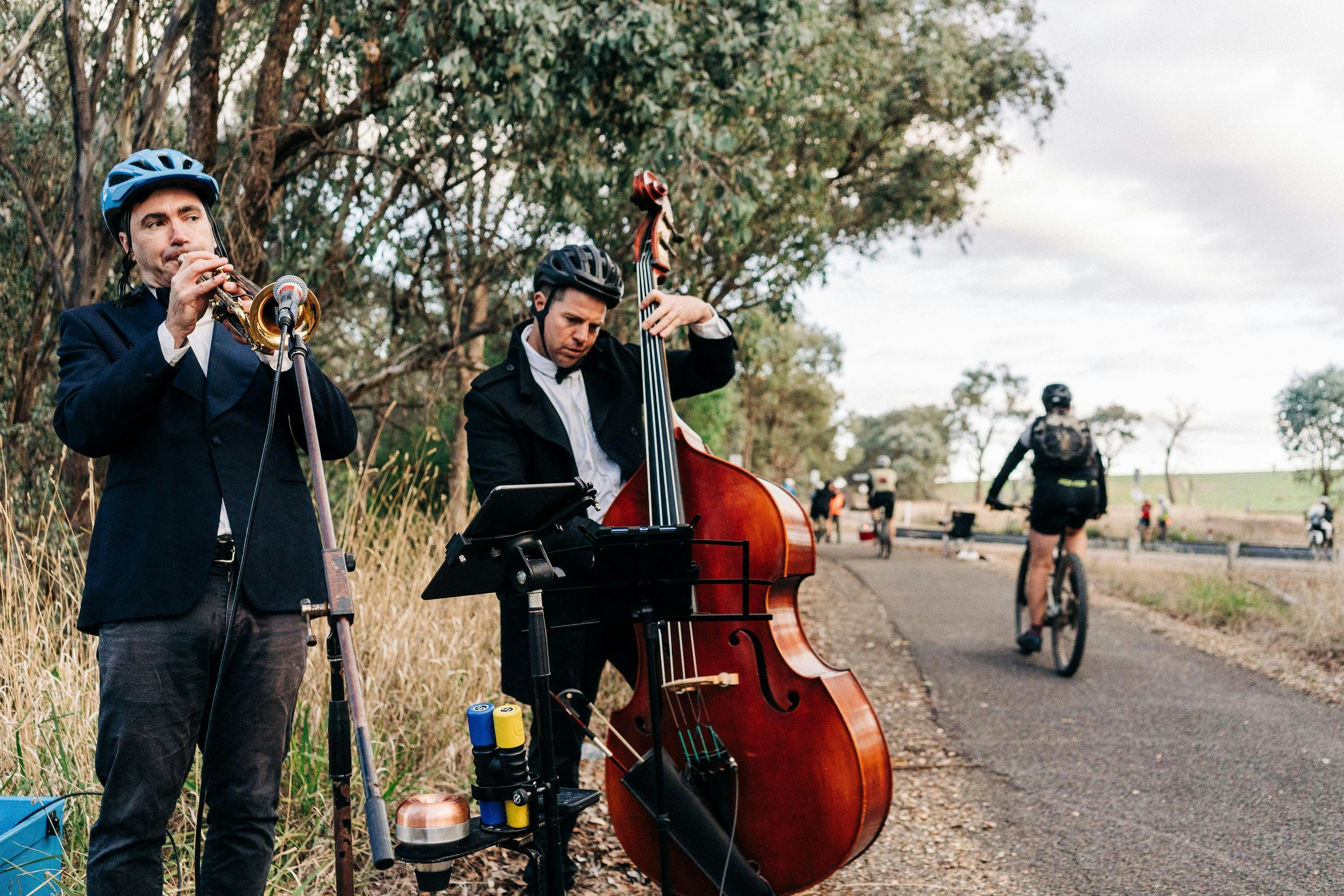 Musicians playing trumpmet and cello while cyclists ride past on bike path
