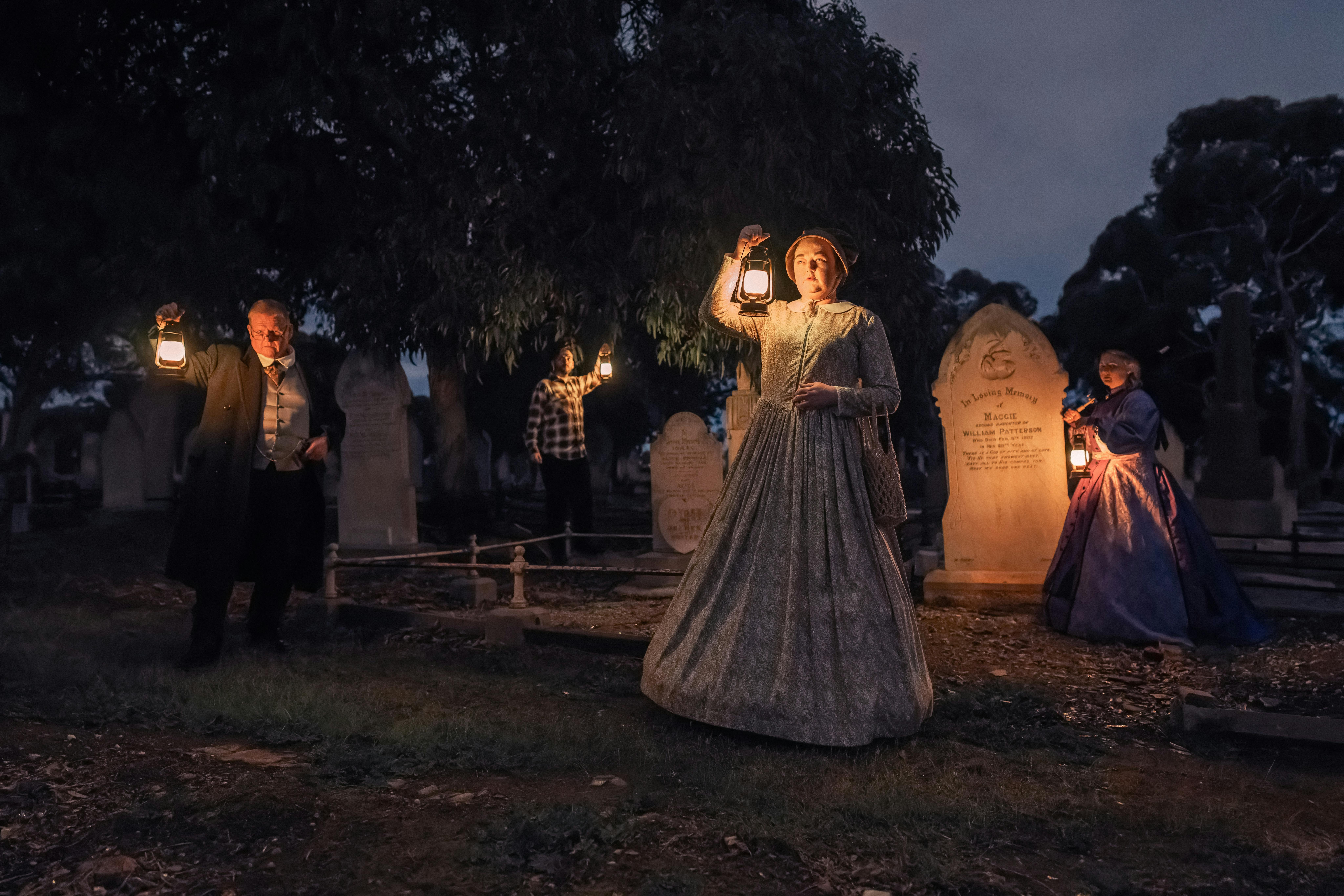 Nighttime in a cemetery, 5 people dressed in 1900's clothing lit by the lanterns they are holding.