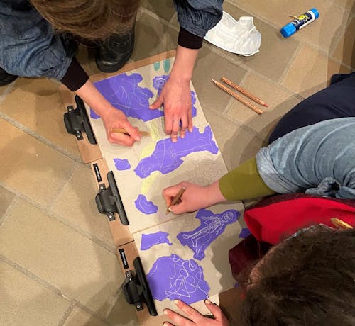 An overhead photograph of two people drawing with pencils on brown and purple paper