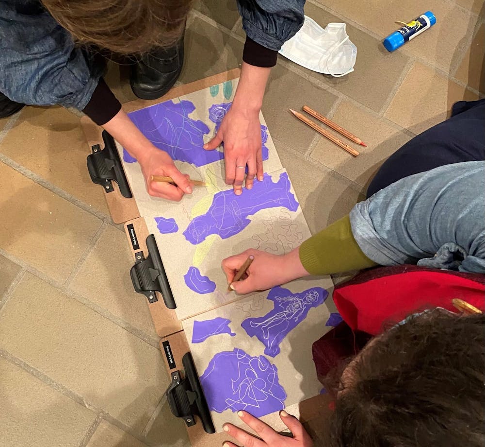 An overhead photograph of two people drawing with pencils on brown and purple paper