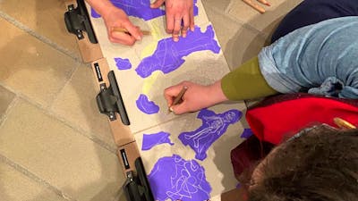 An overhead photograph of two people drawing with pencils on brown and purple paper