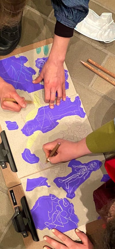 An overhead photograph of two people drawing with pencils on brown and purple paper