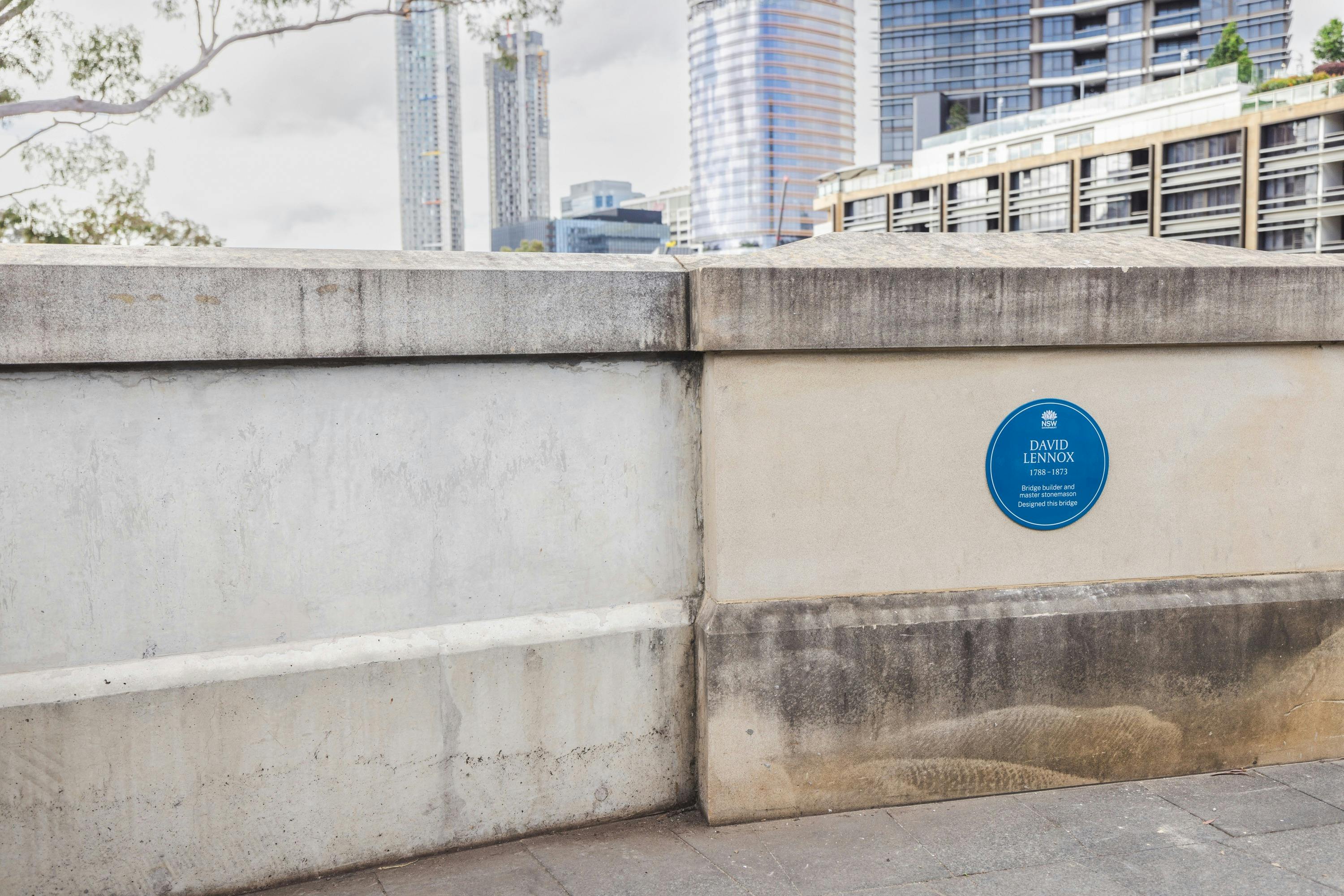 Blue plaque on low concrete barrier of bridge, tall office buildings in the background