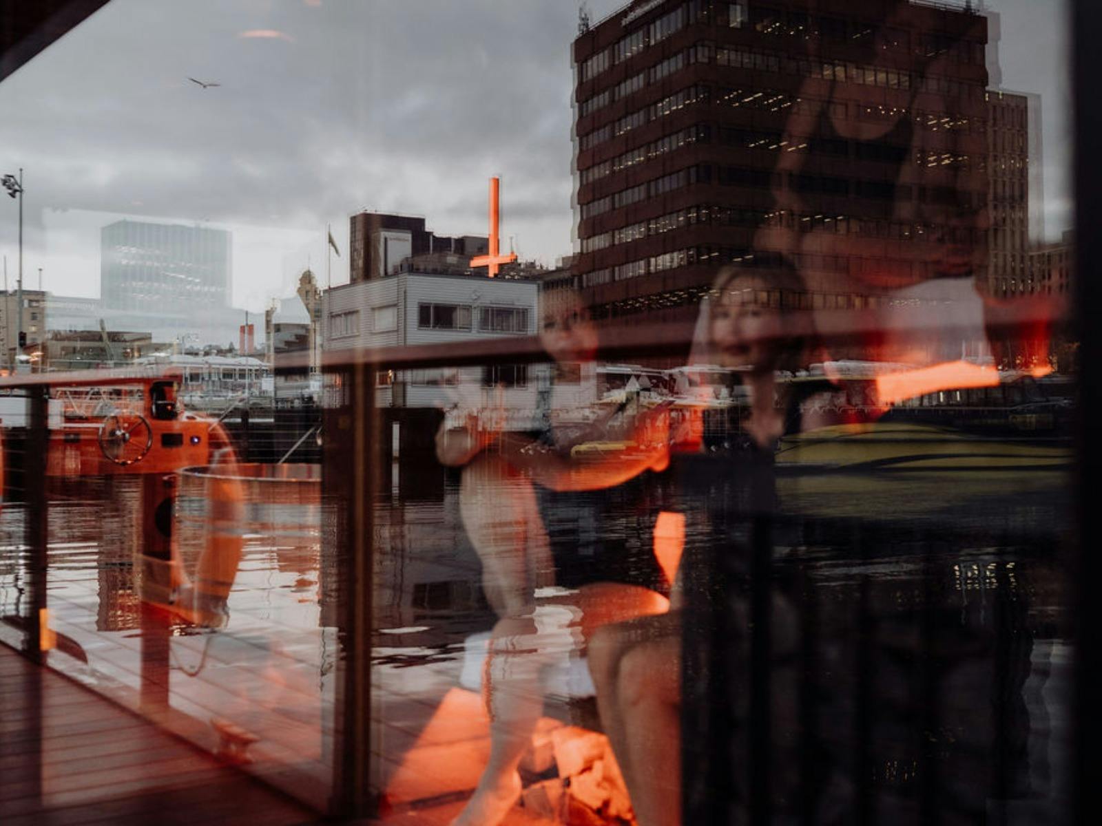 Reflective shot of a sauna interior layered over a harbor and a neon red cross.