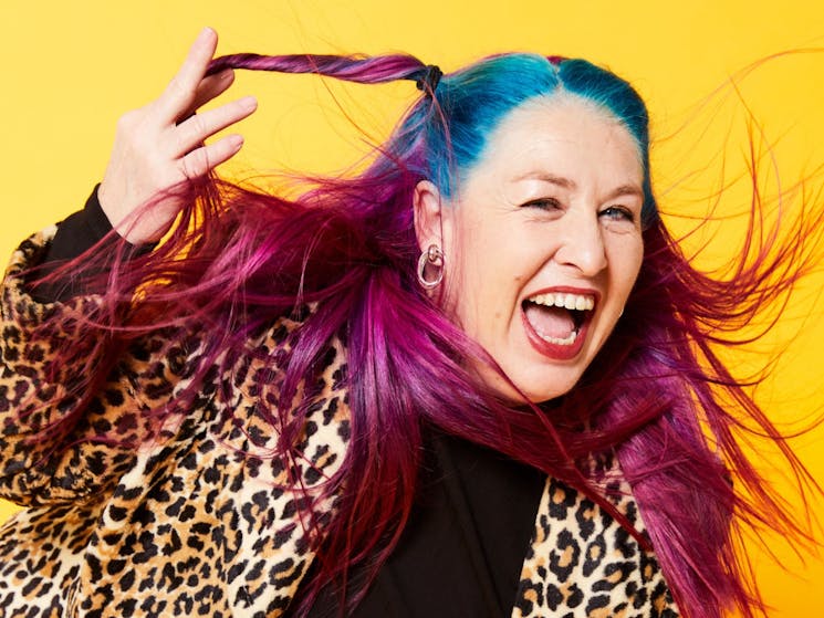 Kelly Mac looks off camera and laughs. She is a middle-aged Caucasian woman with pink and blue hair.
