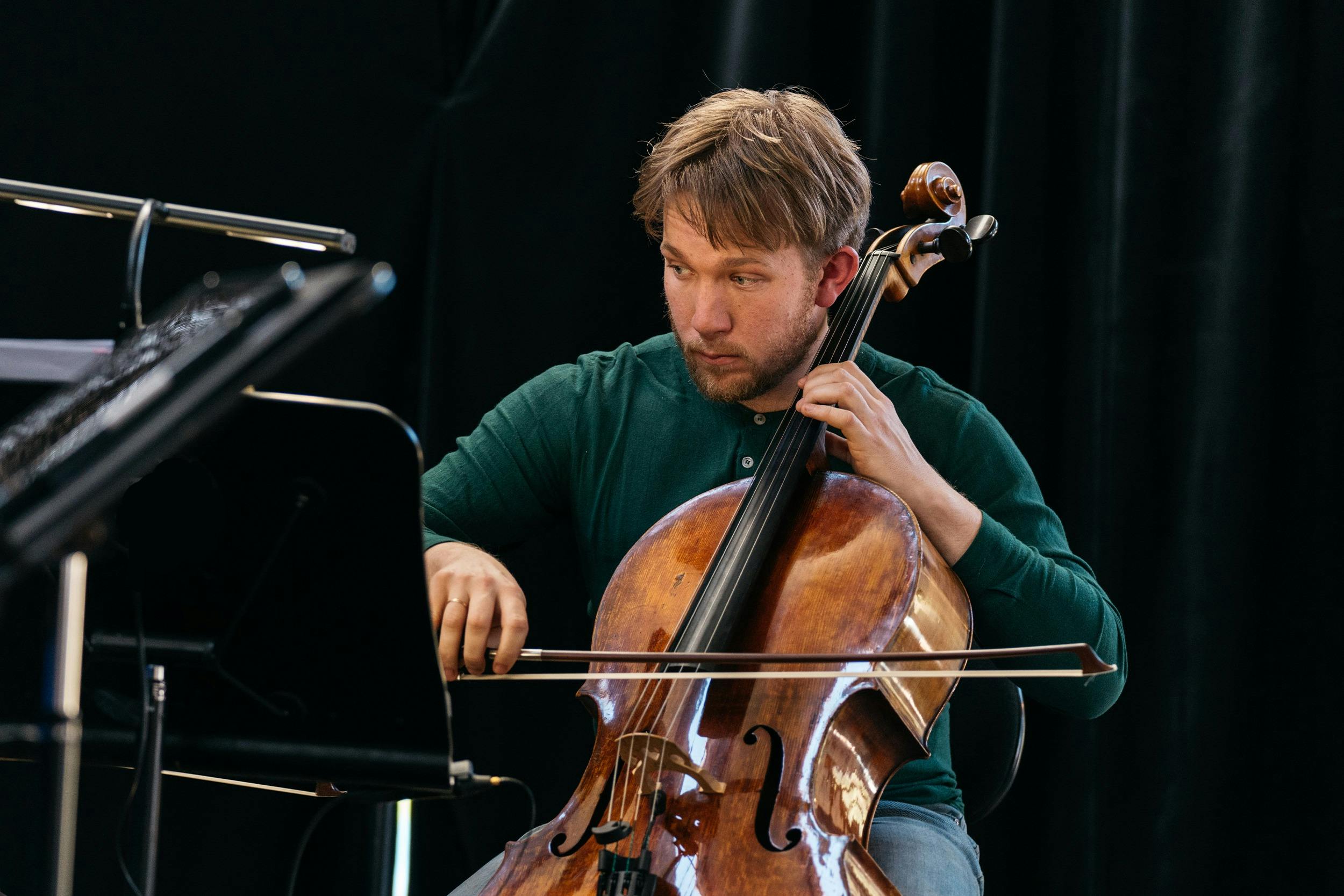 Jonathan Békés seated and playing a cello, holding the bow in the right hand and pressing the string