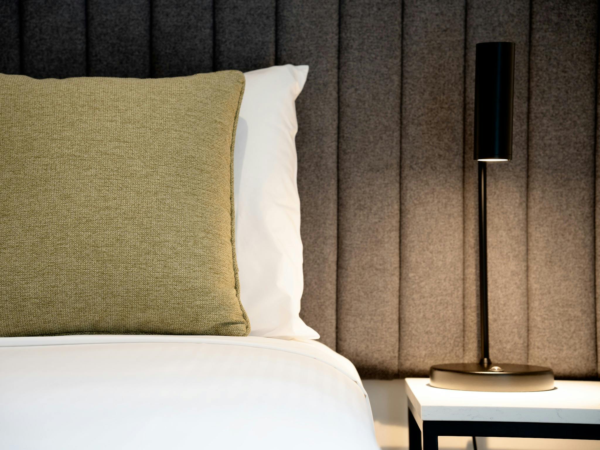 Close up shot of a bed with upholstered headboard. A lamp illuminates the marble bedside table.