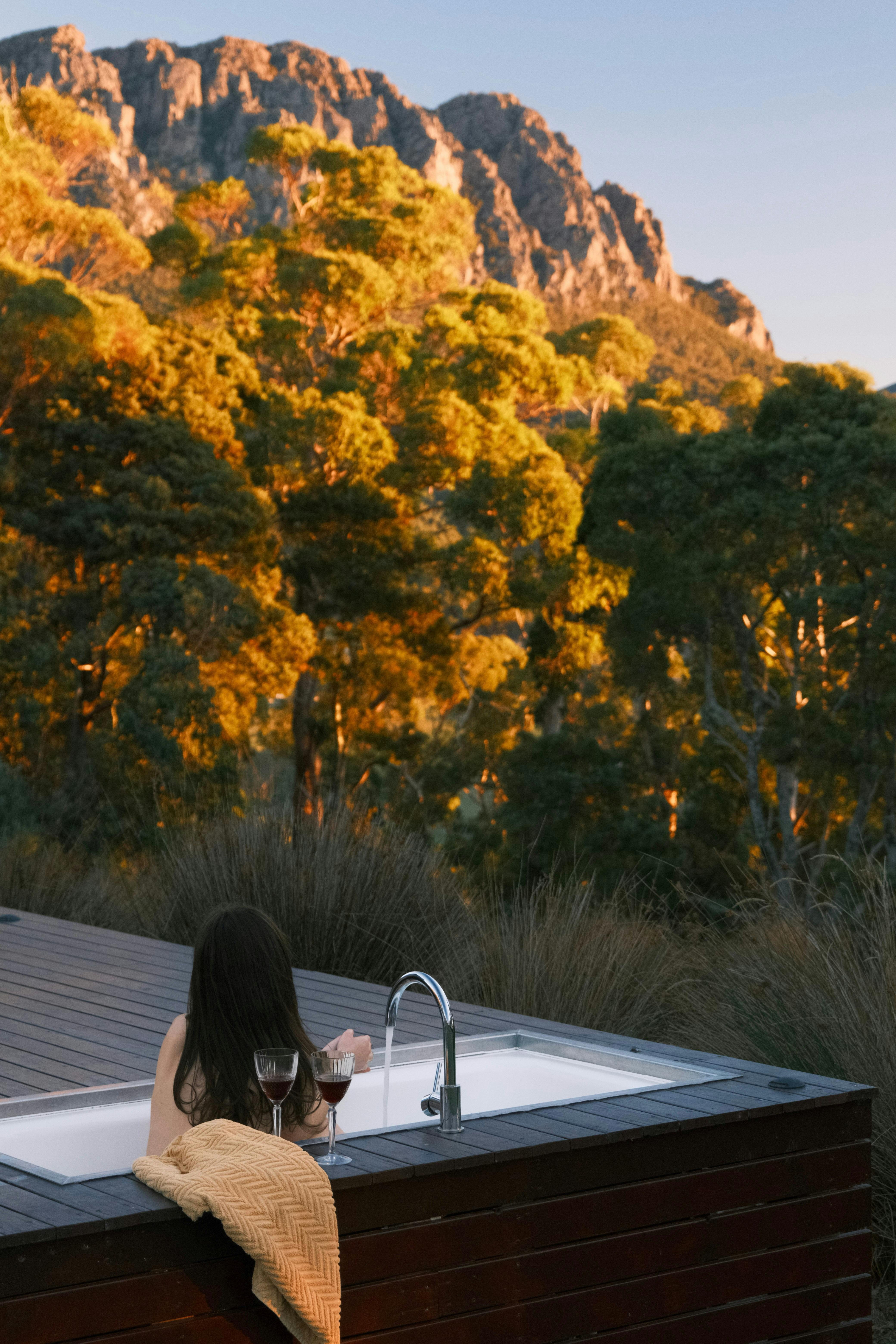 A sunken deck bath with a dark haired woman looking at the mountain with setting sun