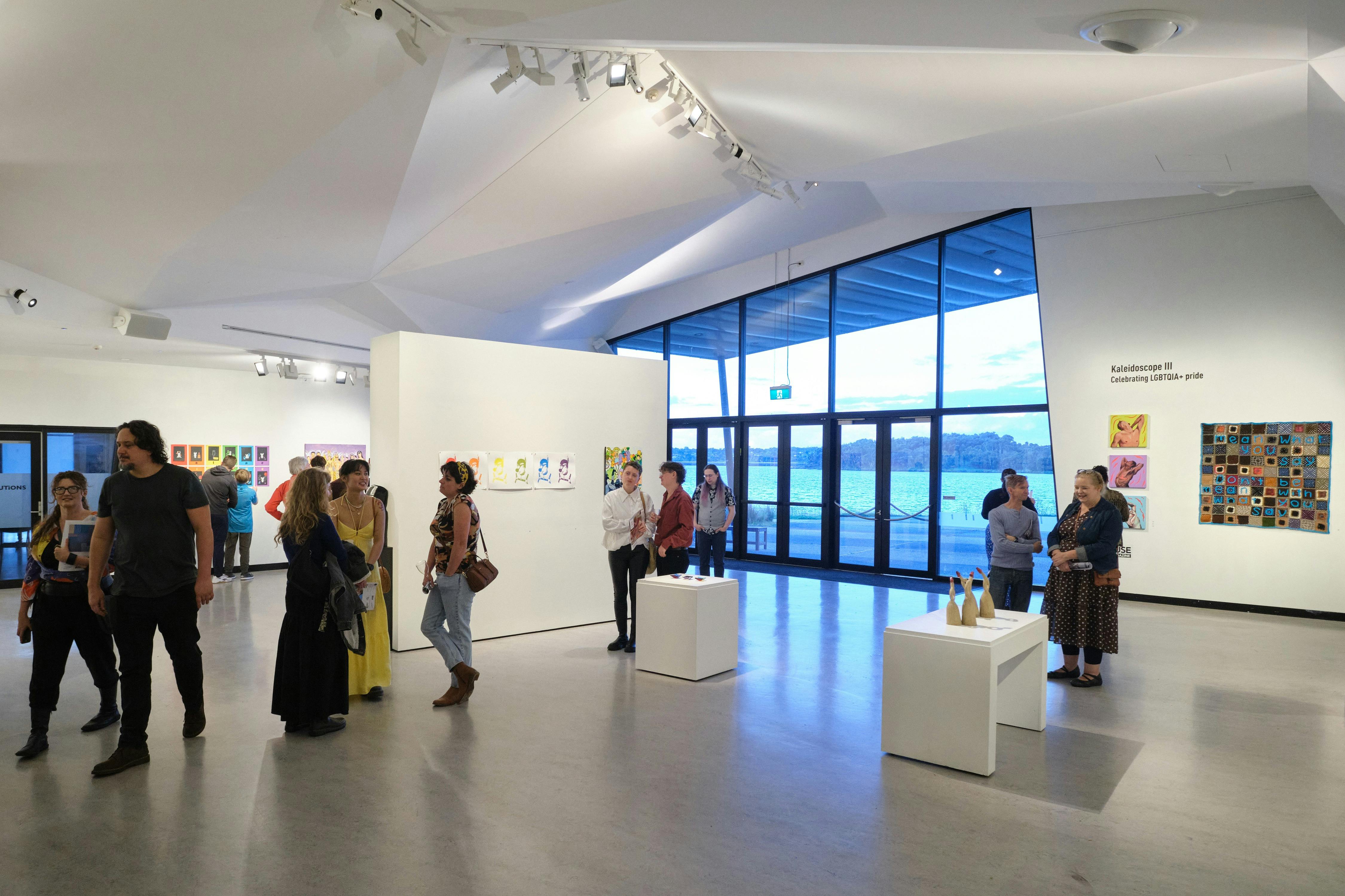 People attend an exhibition in a spacious gallery, with large windows looking on to the lake.