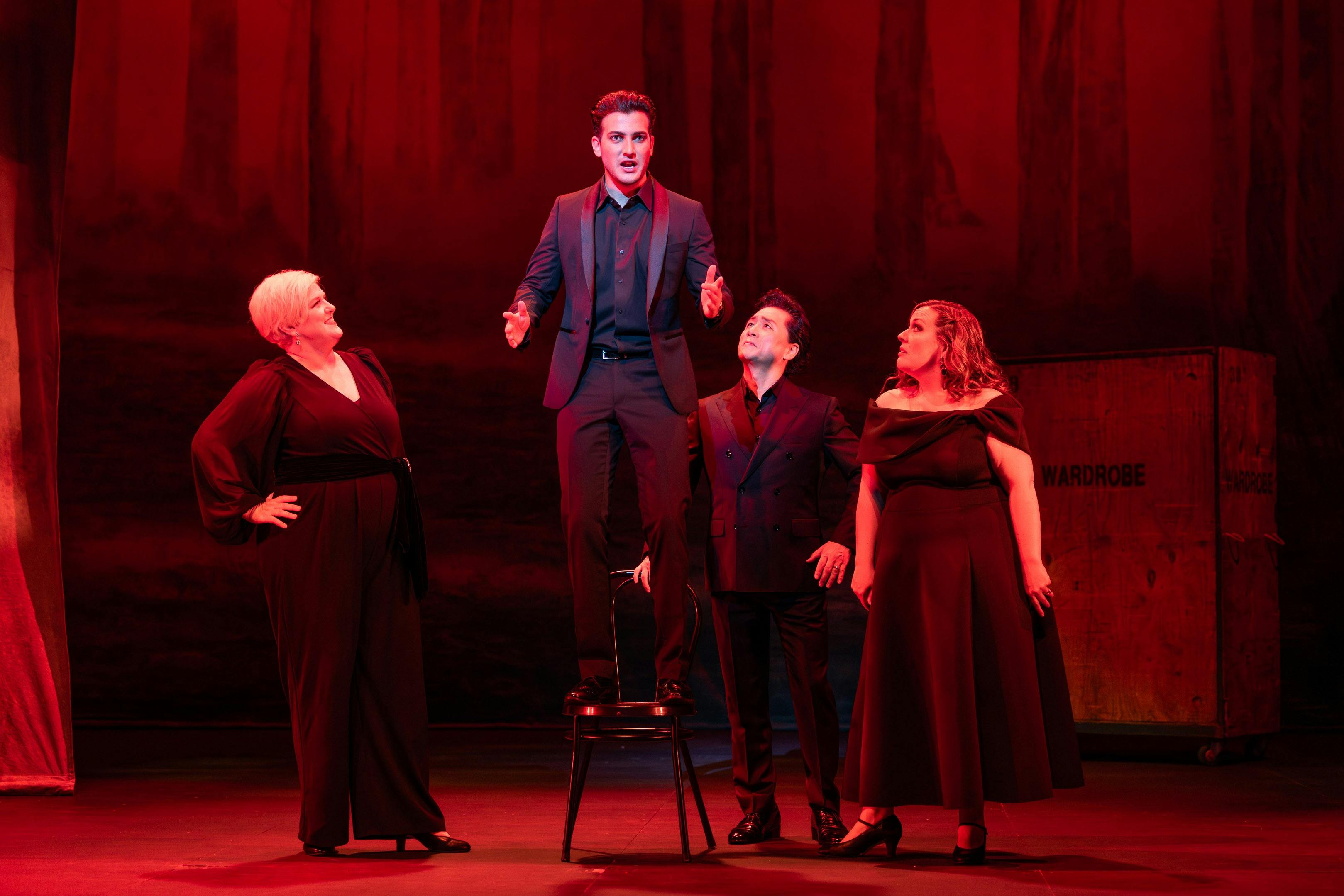 3 Opera singers stand around a baritone in a suit standing on a chair singing
