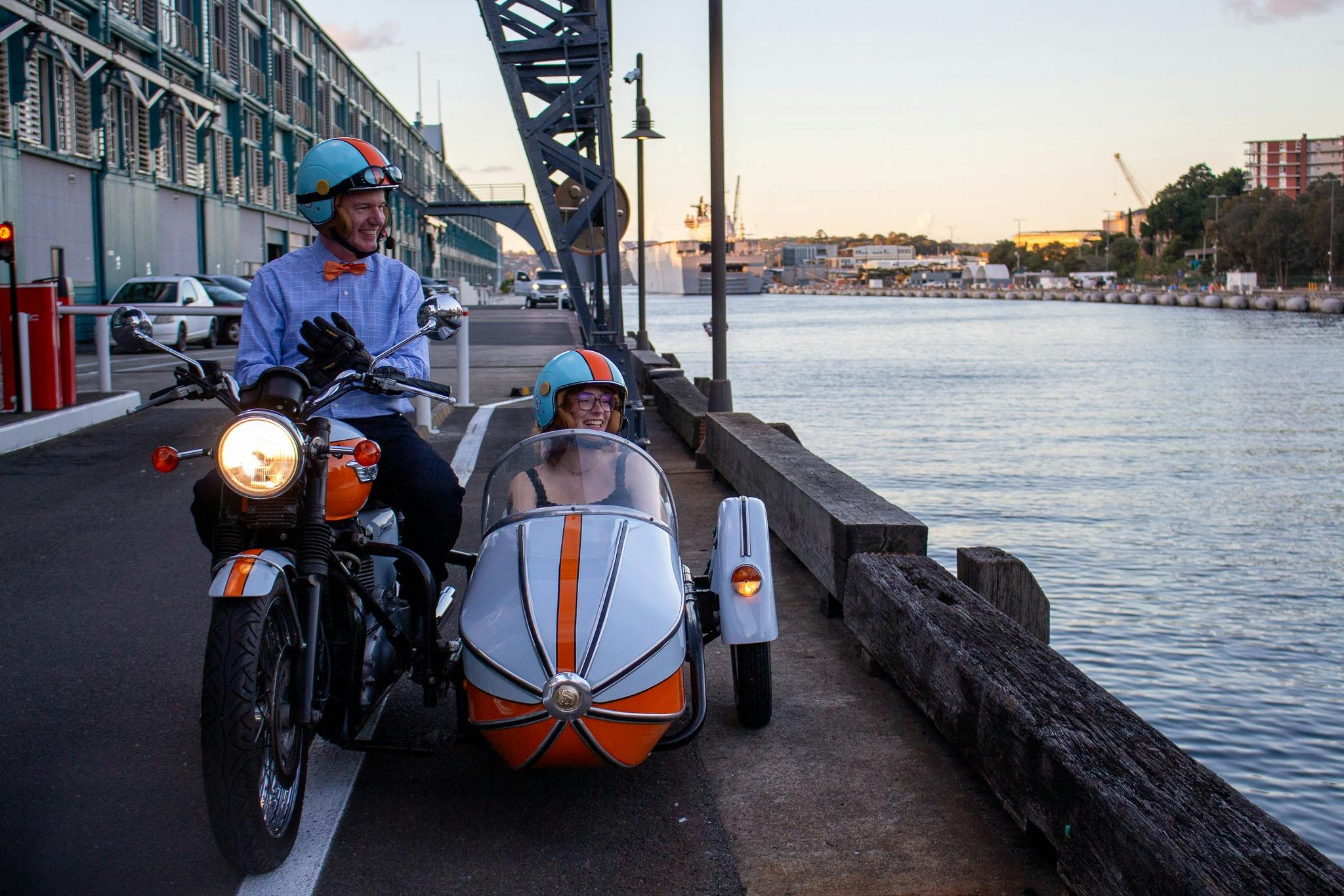 A motorcycle and sidecar by Sydney Harbour with smiling passengers at dusk