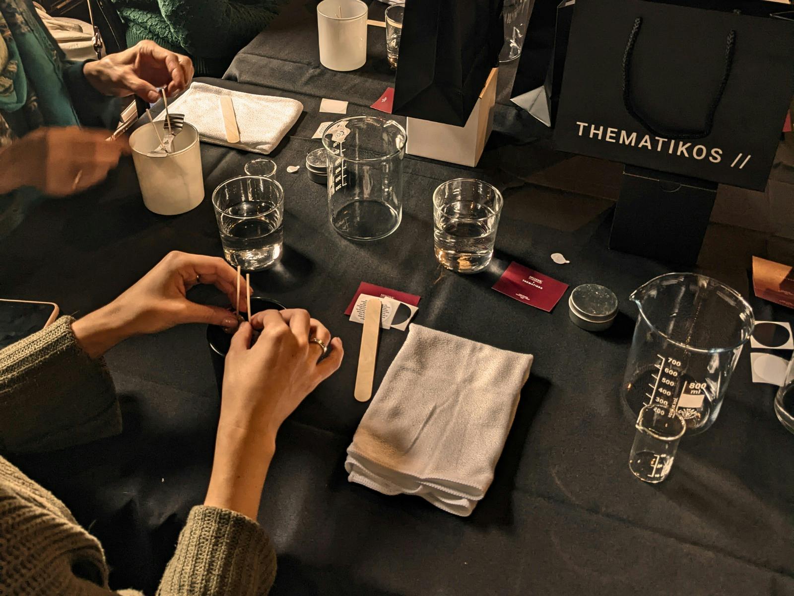 A workshop table with candle making materials and a pair of hands placing wicks inside a jar