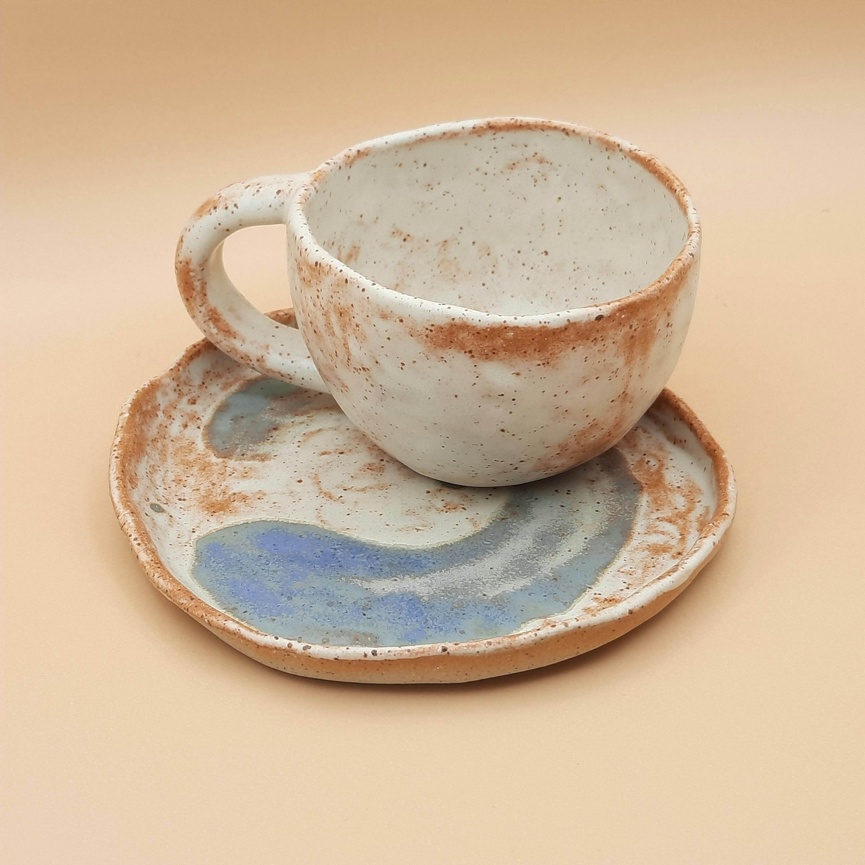 A handmade ceramic cup sitting upon a matching plate, with white and earthy colours and a blue swirl