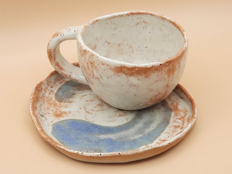 A handmade ceramic cup sitting upon a matching plate, with white and earthy colours and a blue swirl