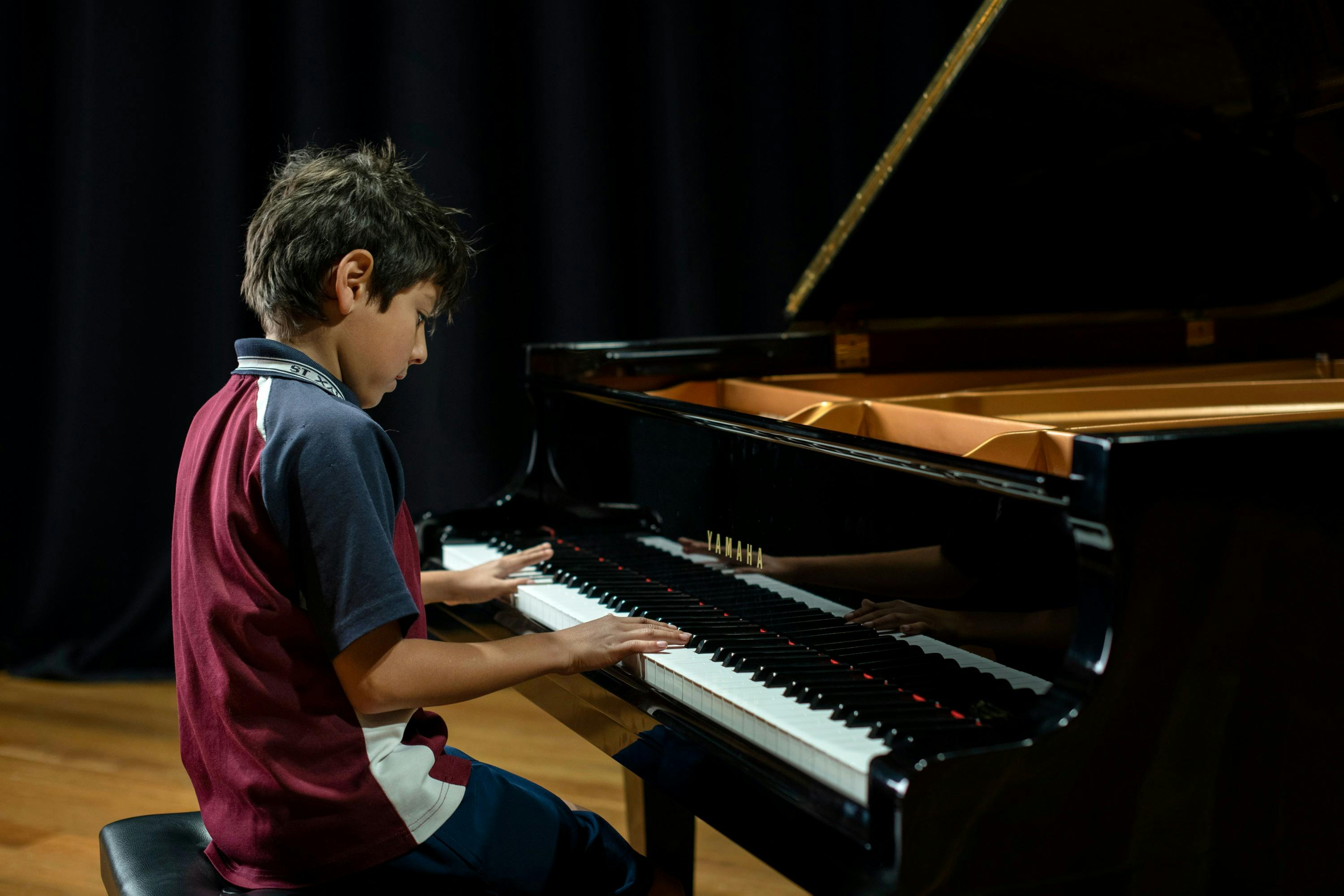 Ayoung boy in a maroon and blue polo shirt sits at a Grand Piano