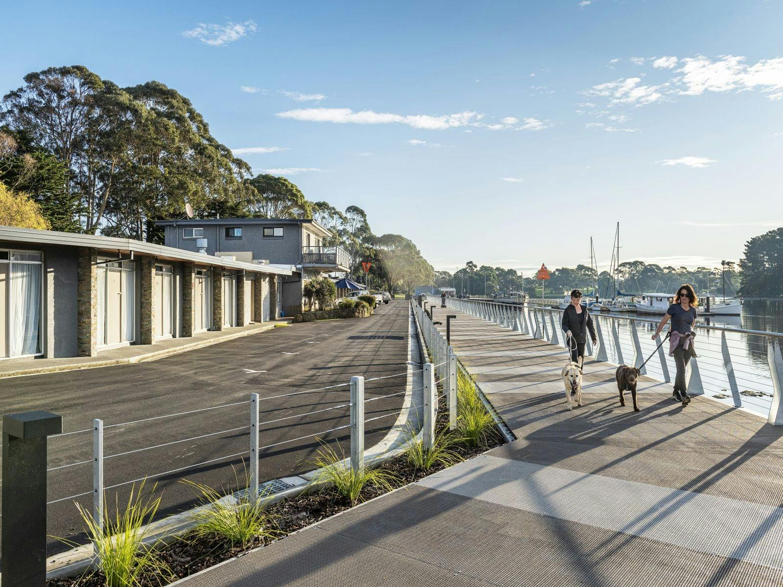 Two people walking their dogs along a modern boardwalk along the  Inglis river motel on left