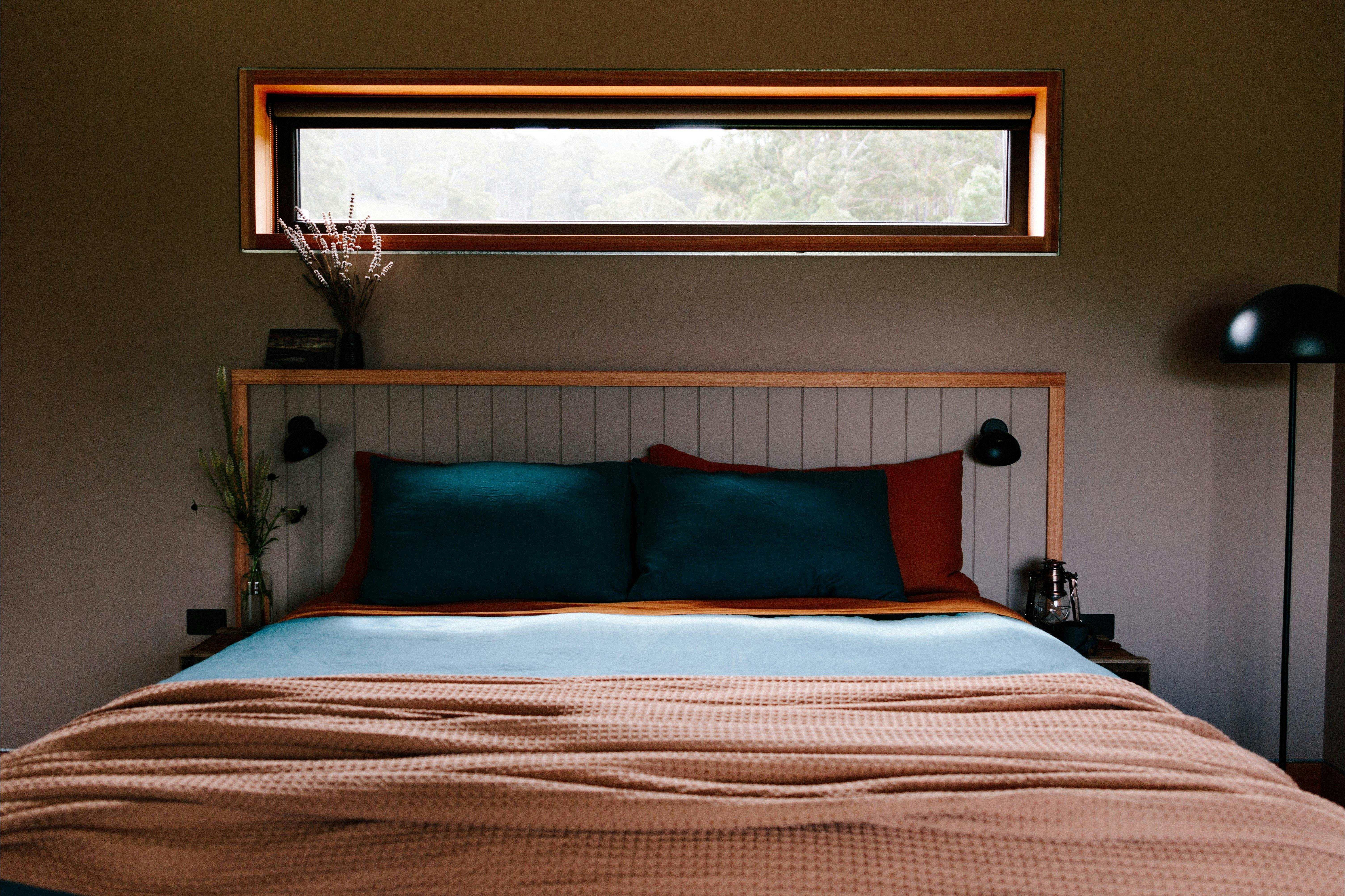 A bed with rust and blue linen and a throw rug. There's a window above the bed and a floor lamp.