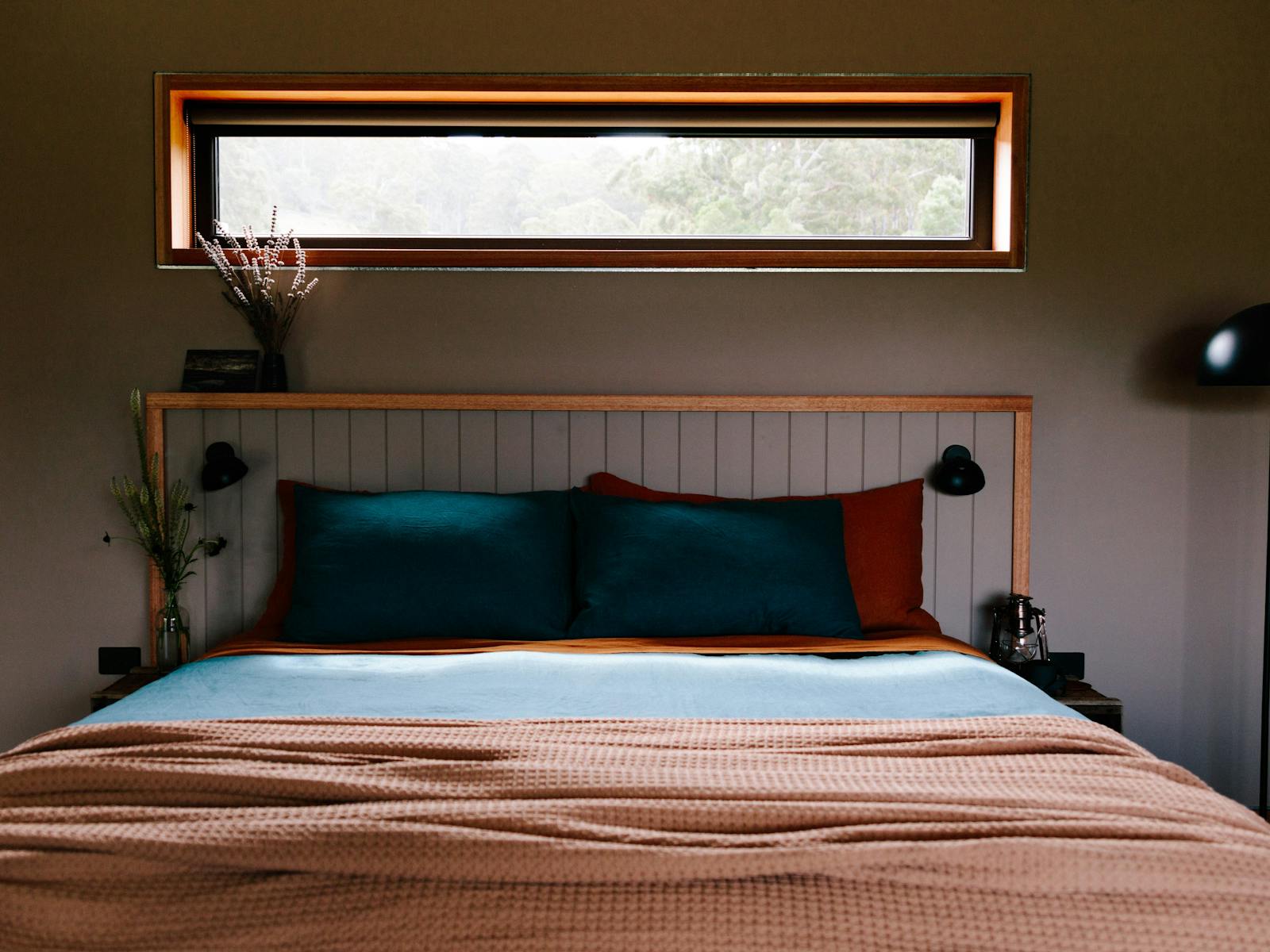 A bed with rust and blue linen and a throw rug. There's a window above the bed and a floor lamp.