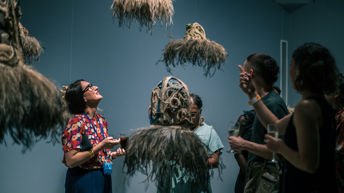 A group of people stand in the gallery surrounded by hanging woven works with emu features.