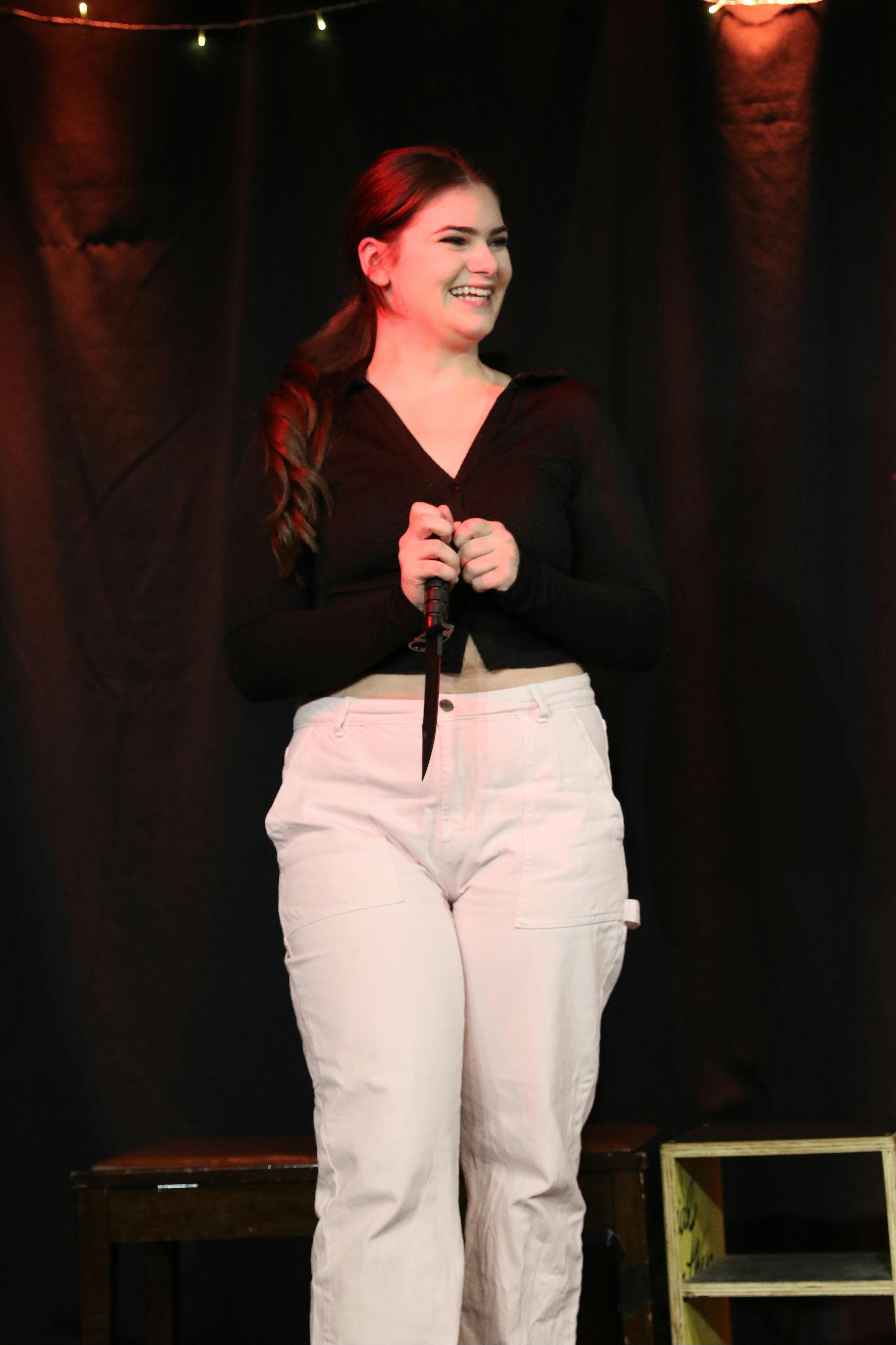 A young woman, standing on a stage, laughs, holding a bowie knife in her hand.