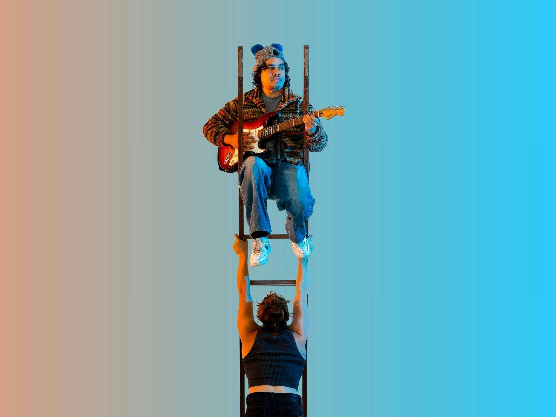 A guy in a beanie playing guitar on a ladder that is being held up by a muscular acrobatic woman
